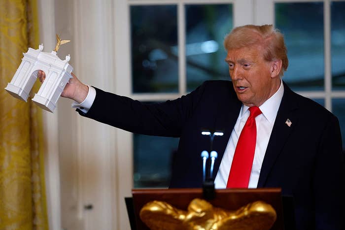 A person in a dark suit and red tie holds a miniature white arch model during a speaking event, standing behind a podium