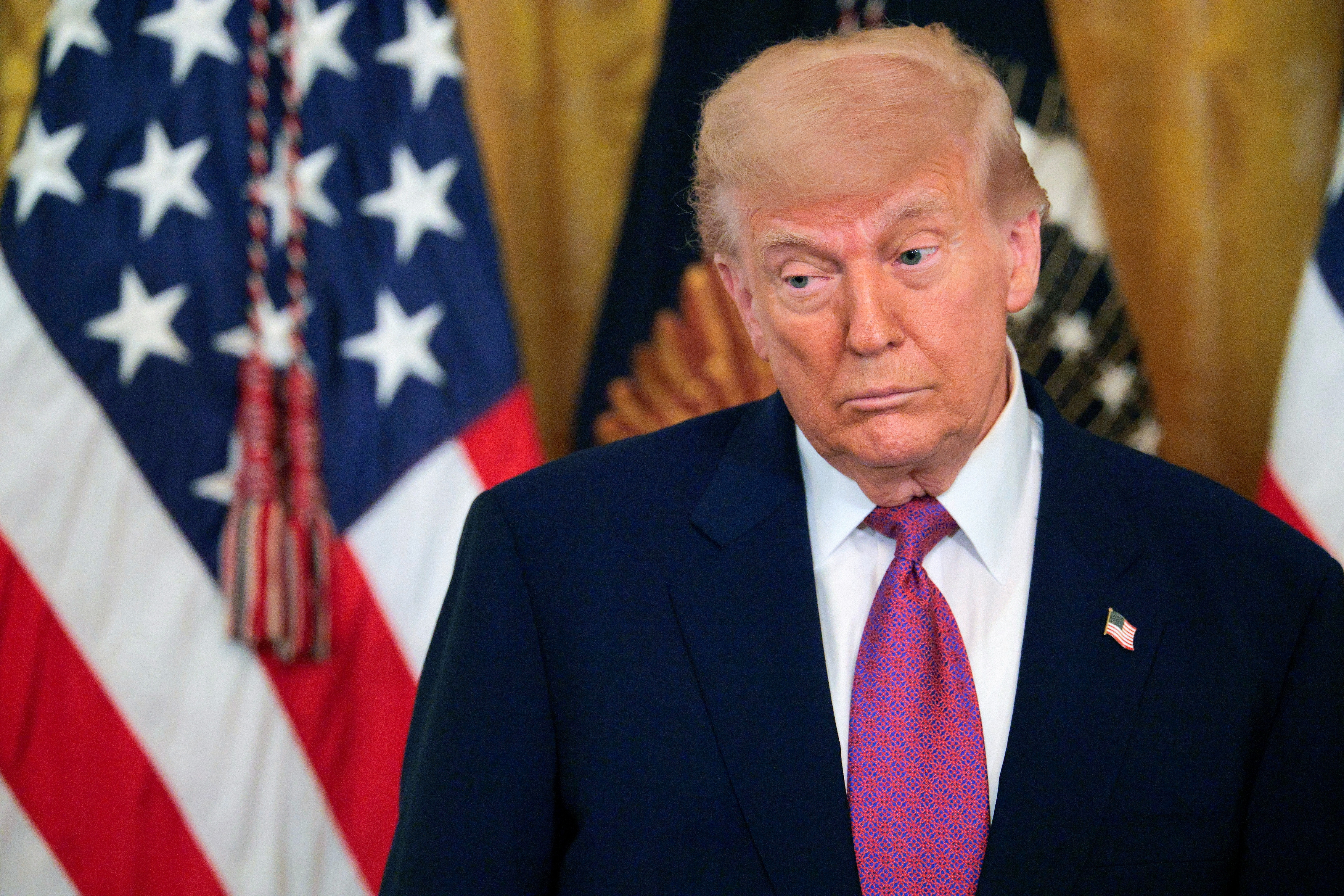 En person i dress og slips står i en formell setting med amerikanske flagg i bakgrunnen