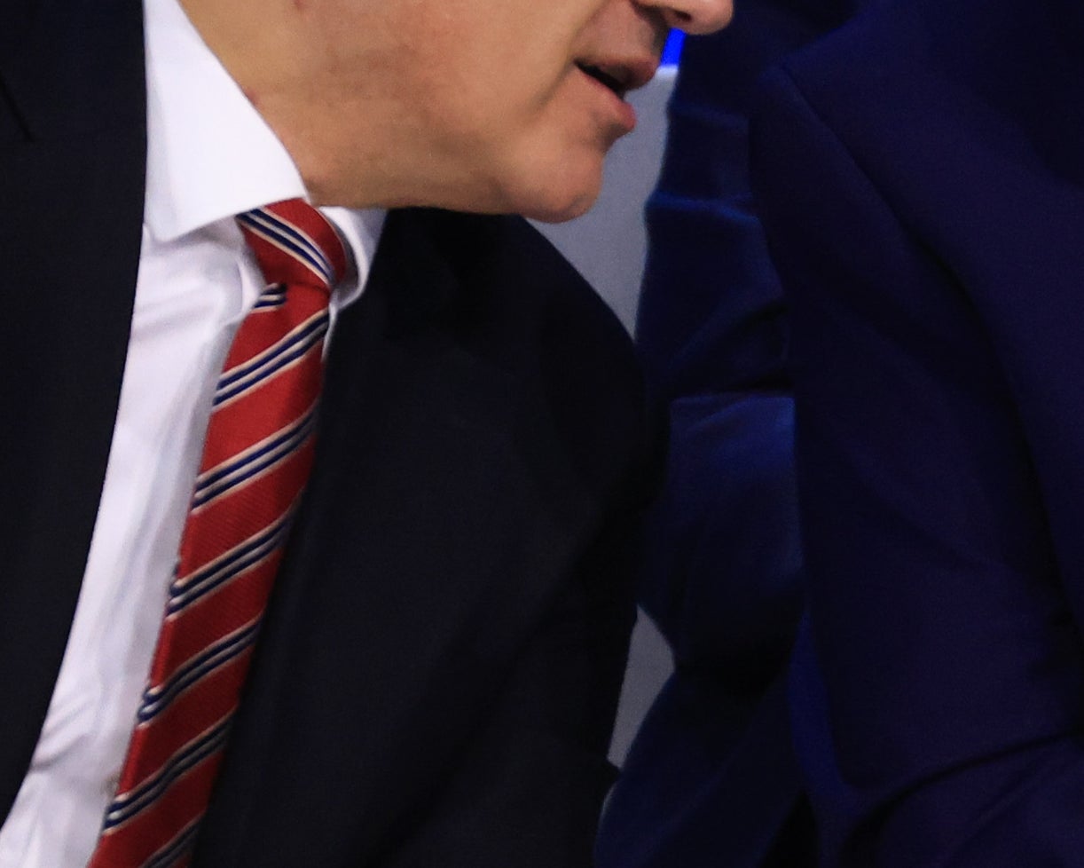 A man in a suit whispers to another suited man seated next to him at a formal event, with others in business attire in the background