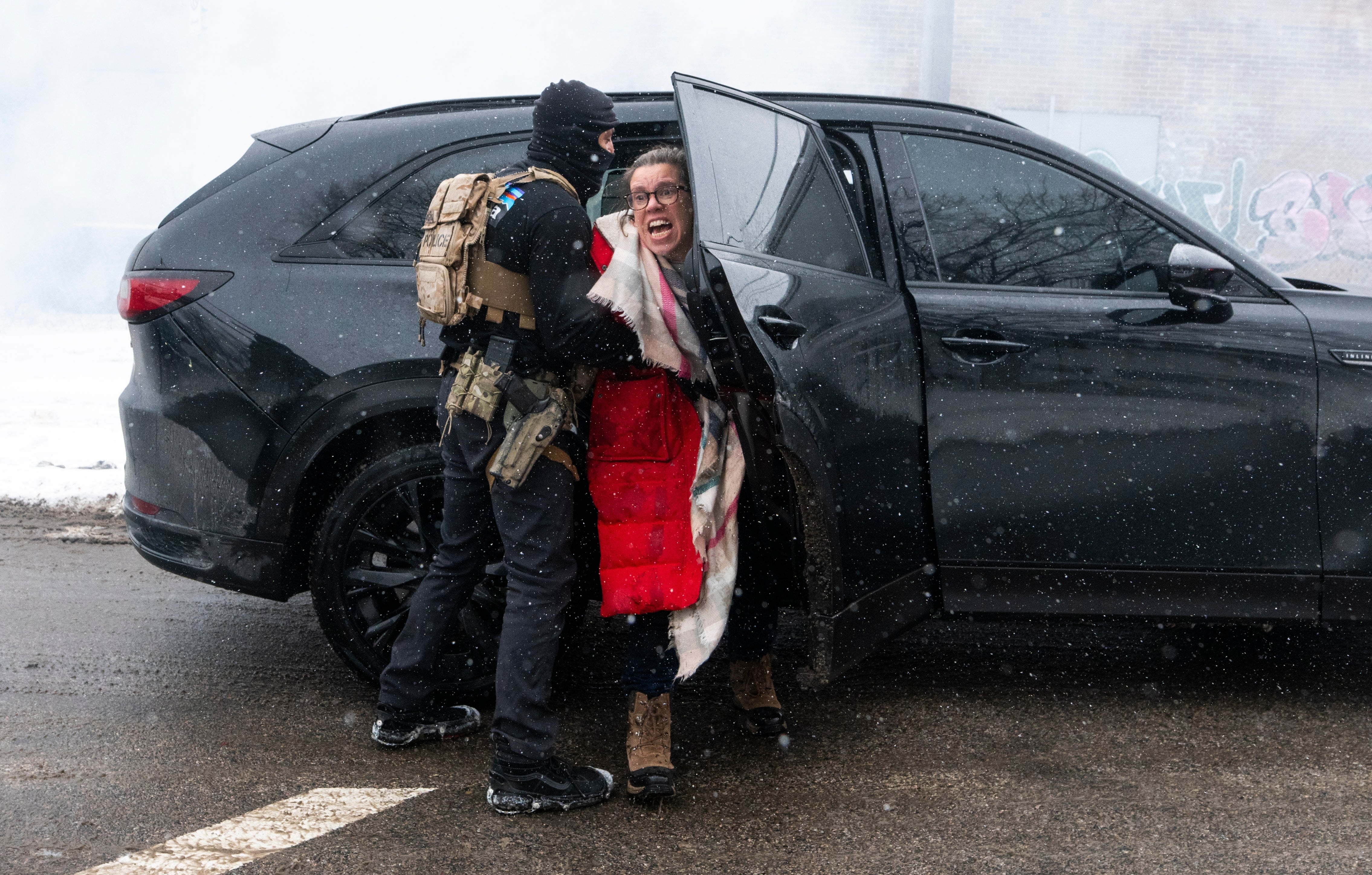 Person i taktisk utstyr som holder en kvinne tilbake utenfor et kjøretøy på en snødekt gate; hun virker synlig opprørt