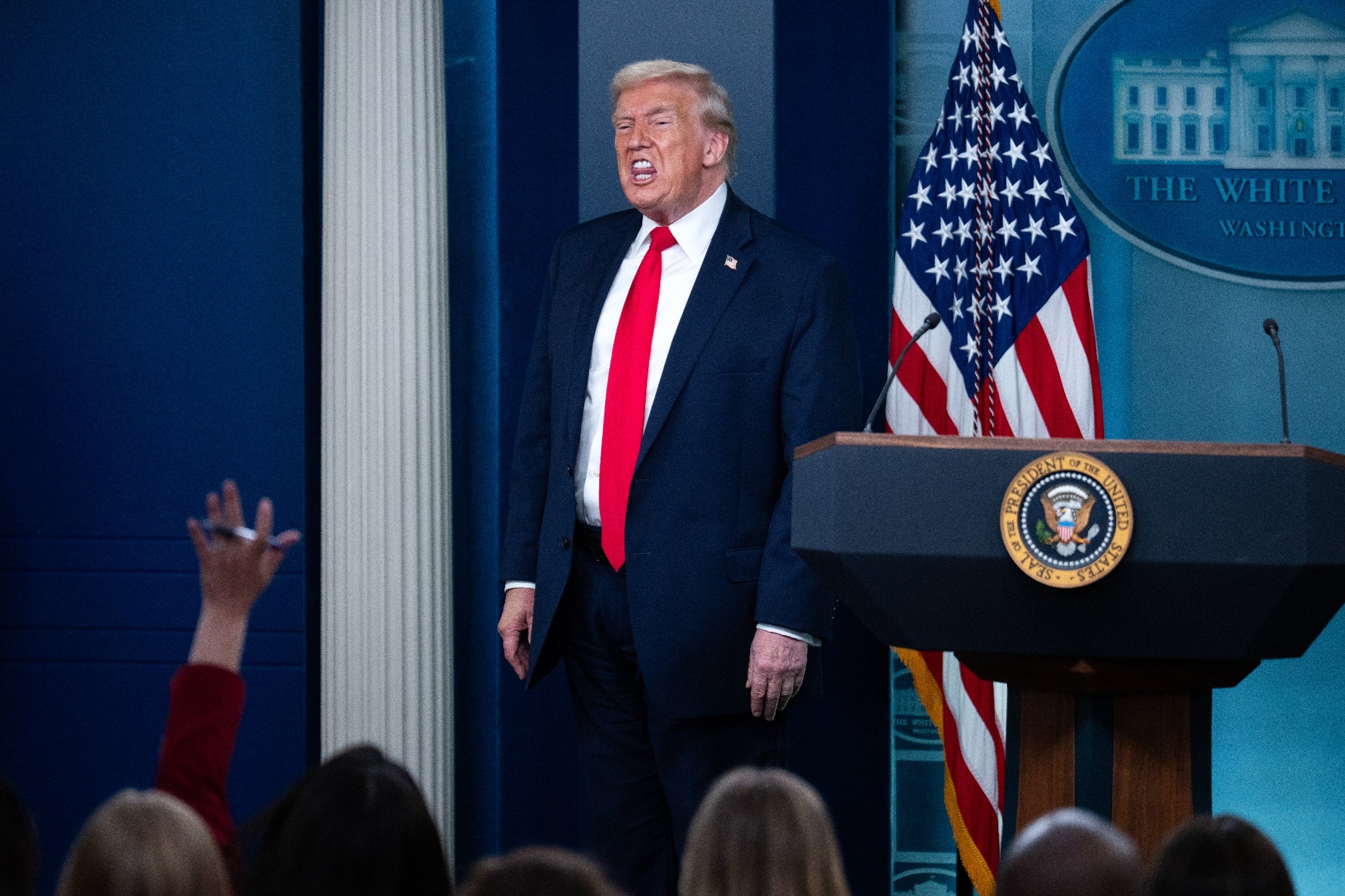 En mann ved et pressepodium i dress med rødt slips snakker til journalister, som rekker opp hendene for å stille spørsmål