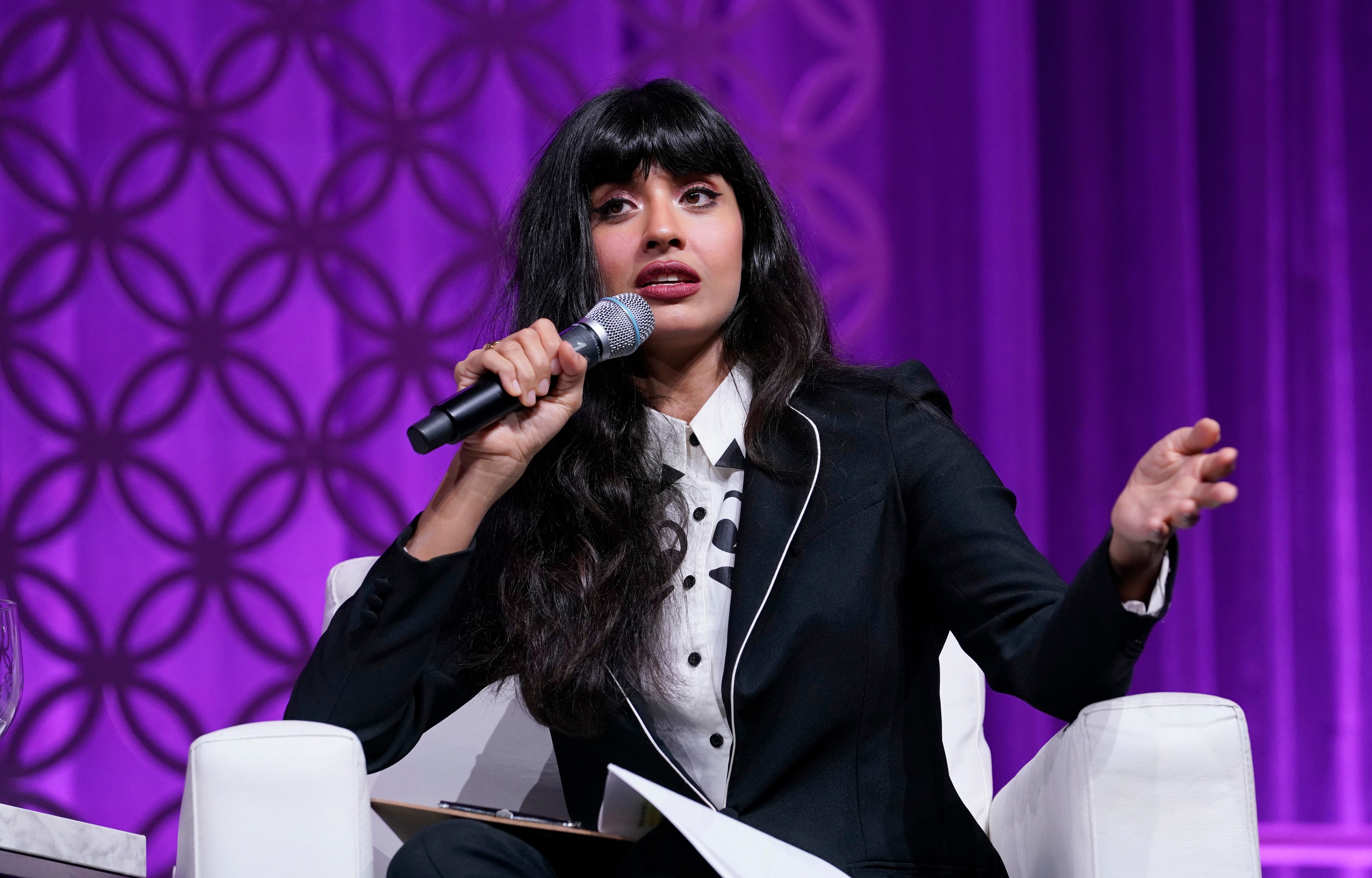 Jameela Jamil in a stylish suit with a polka dot shirt underneath speaks into a microphone while gesturing with one hand on stage