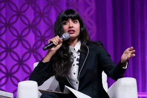 Jameela Jamil in a stylish suit with a polka dot shirt underneath speaks into a microphone while gesturing with one hand on stage