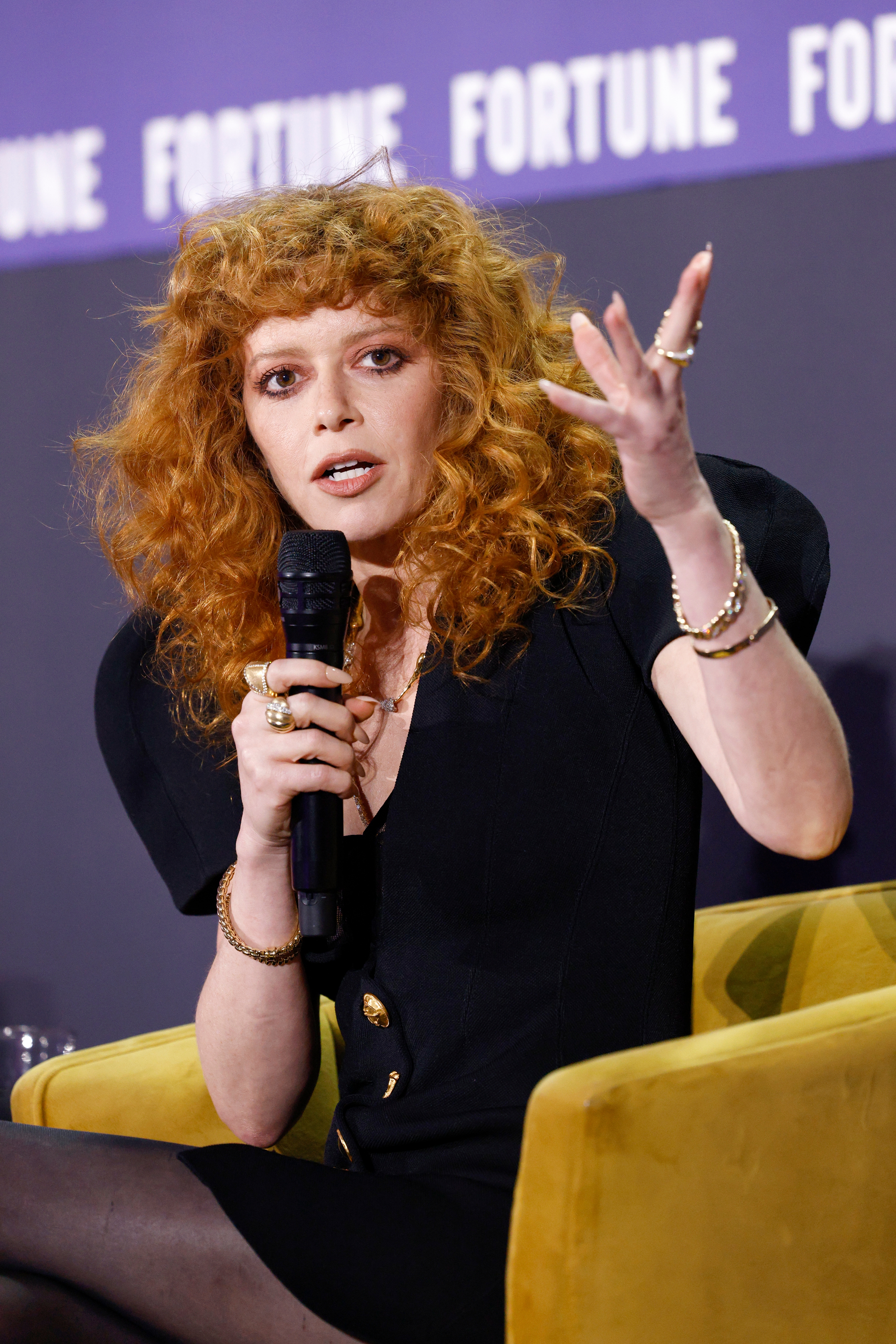Natasha Lyonne med krøllete hår snakker inn i en mikrofon på et panel, iført et stilig antrekk med polstrede skuldre og gulltilbehør