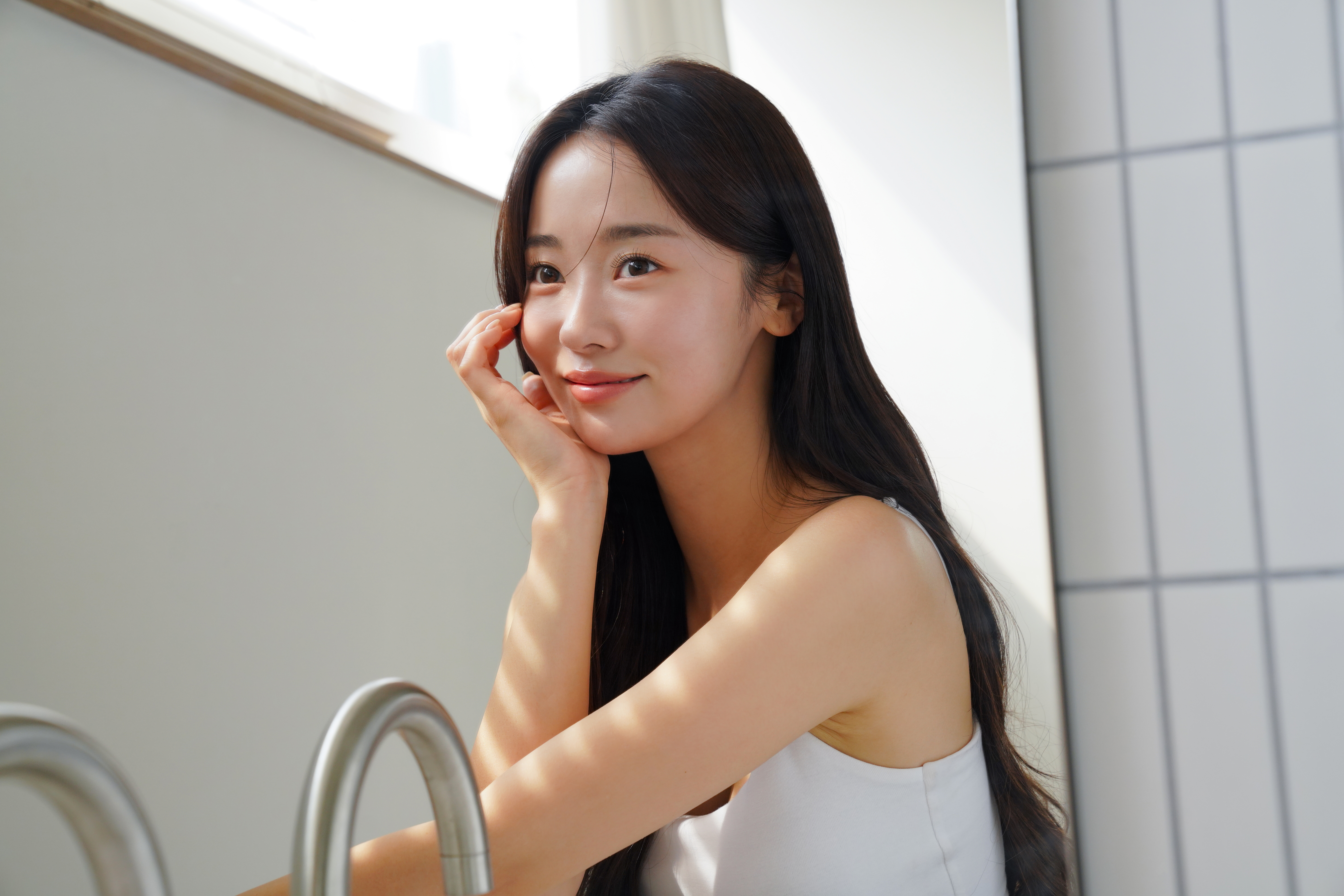 女性が微笑んで窓辺に座っている、カジュアルなタンクトップ姿。リラックスした雰囲気。