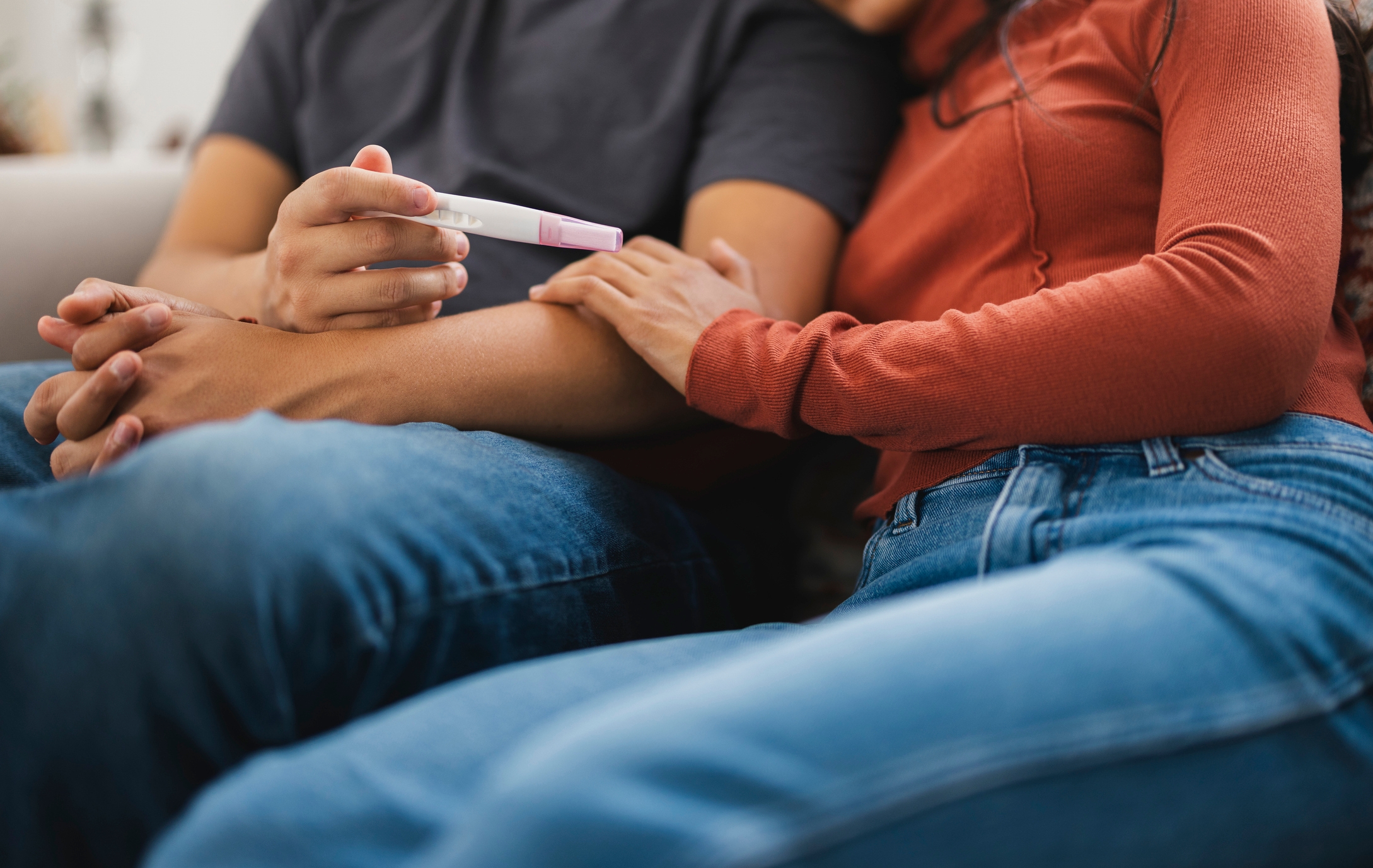 A couple sits closely on a couch, with one holding a positive pregnancy test, signifying a shared, intimate moment