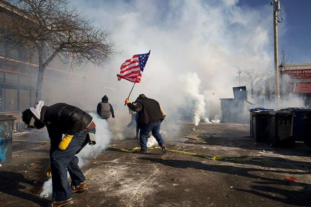 Demonstranter unngår røyk med en person som vifter med et amerikansk flagg blant spredt rusk i urbane omgivelser
