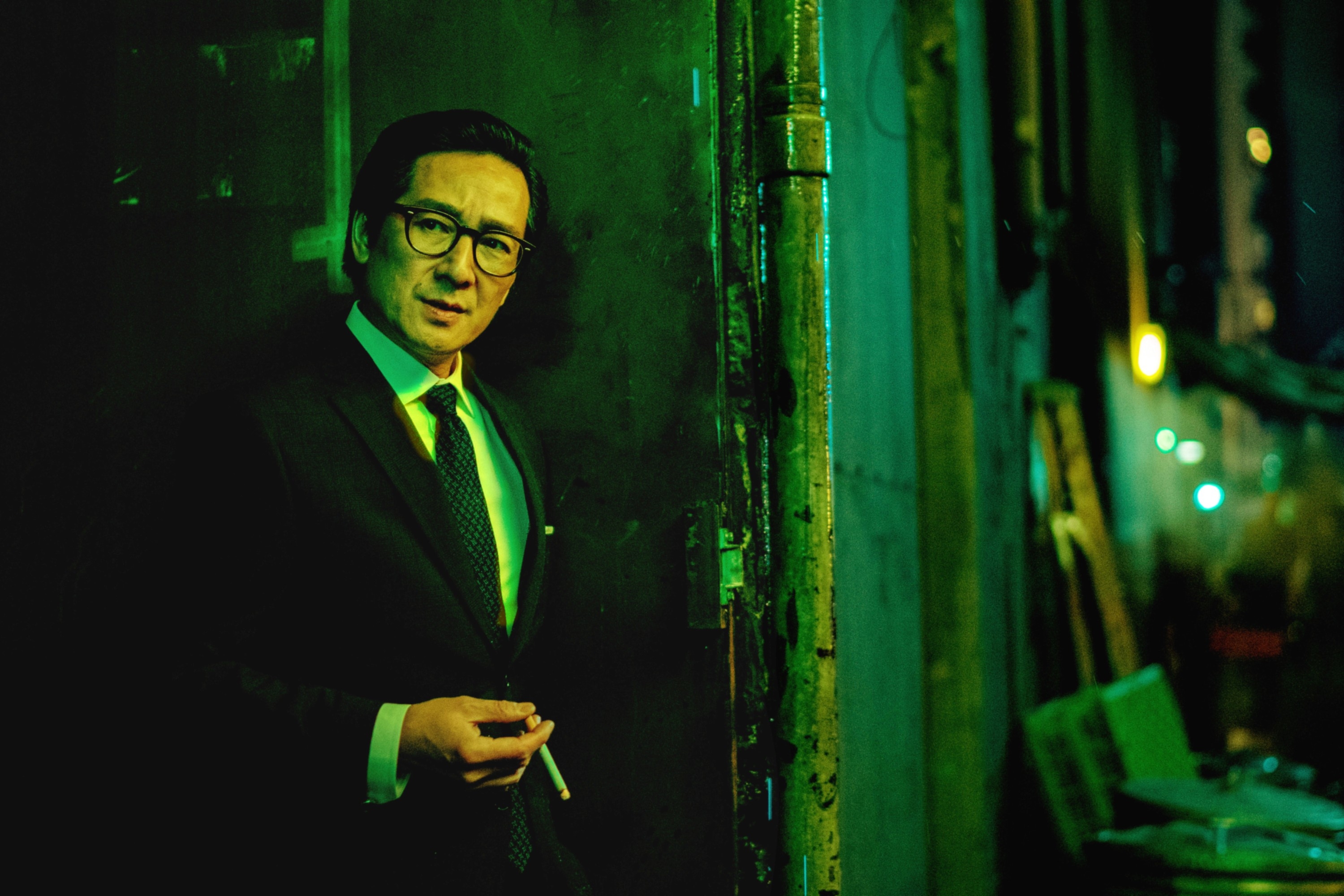 An individual in a suit leans against a wall in a dimly lit alley, holding a cigarette and wearing glasses