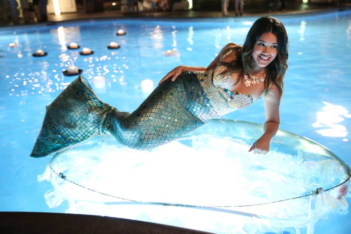 A person dressed as a mermaid poses on a lit platform in a pool, smiling for the camera