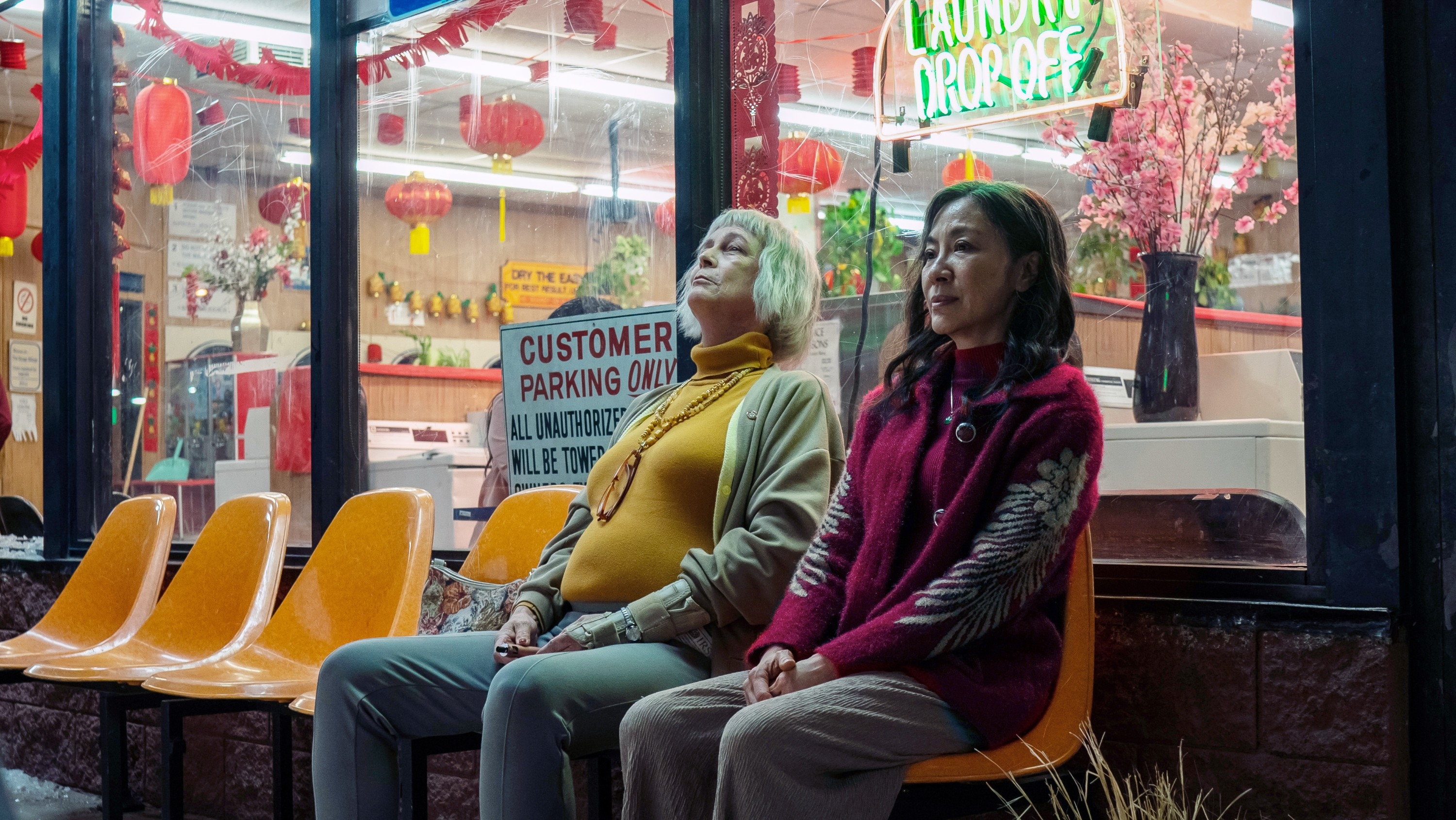 Two people sit on yellow chairs outside a laundromat. One leans back with a relaxed expression, while the other looks thoughtful