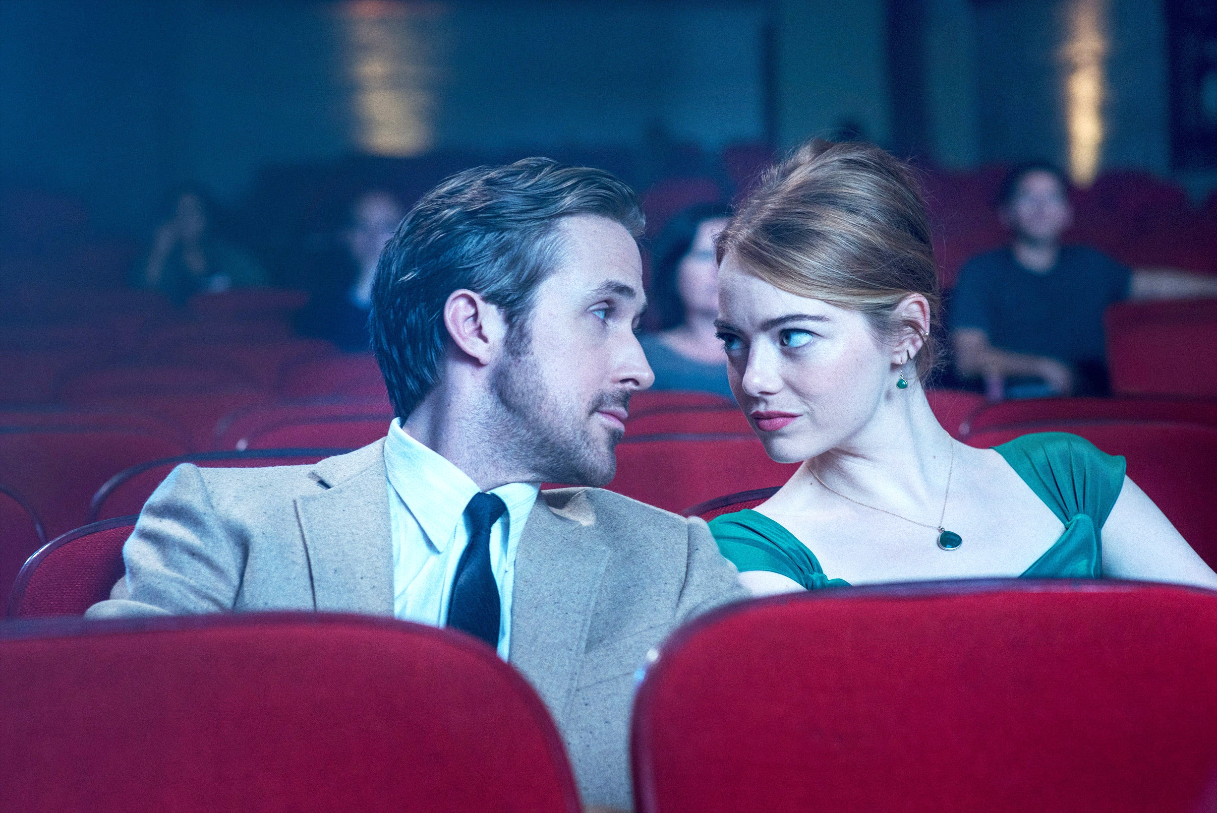 Two people sit in a theater, looking intently at each other. One wears a suit with a tie, and the other a formal dress