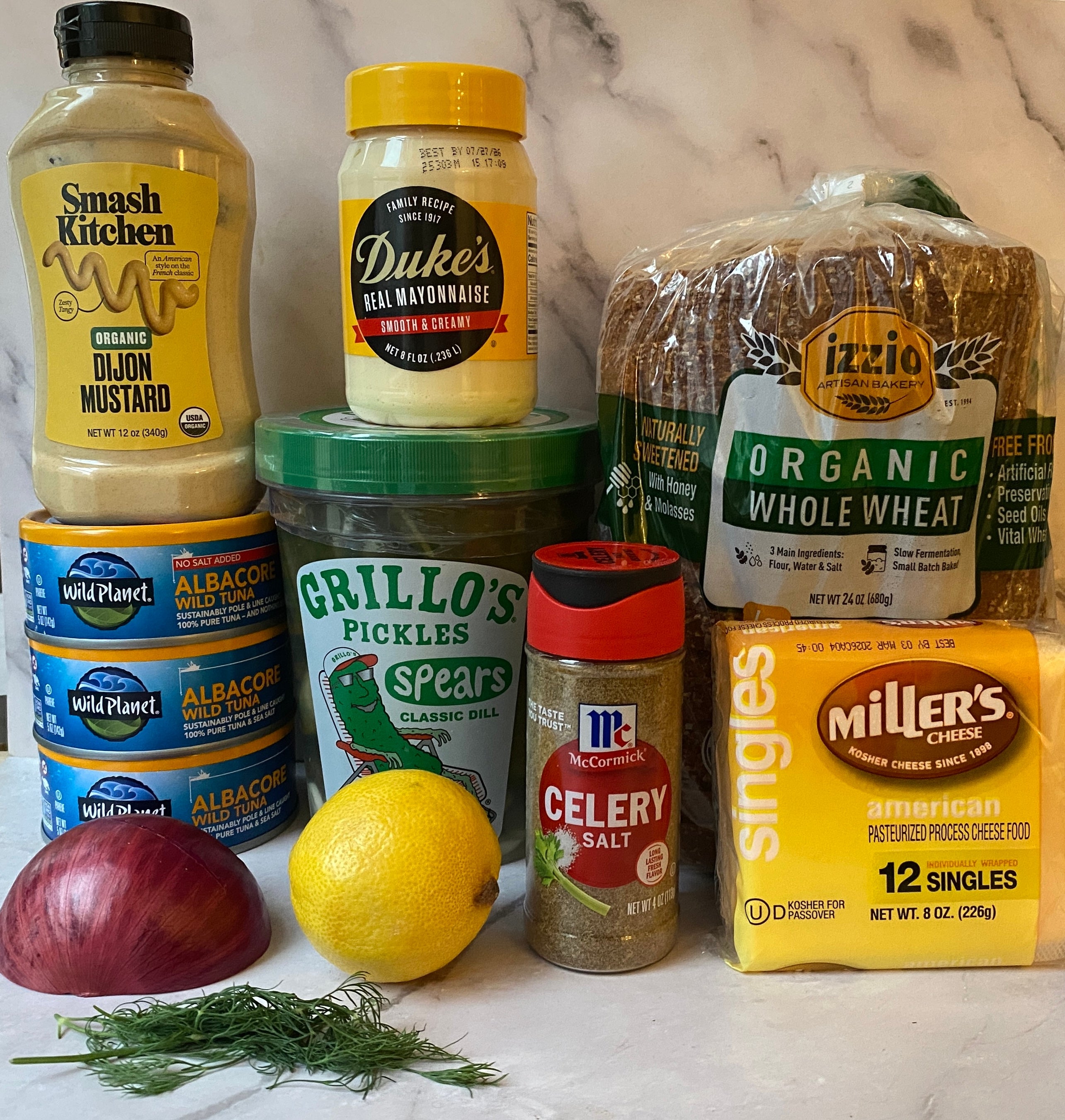Assorted ingredients and products on a countertop: mustard, mayonnaise, pickles, bread, cheese slices, tuna cans, celery salt, onion, lemon, and dill