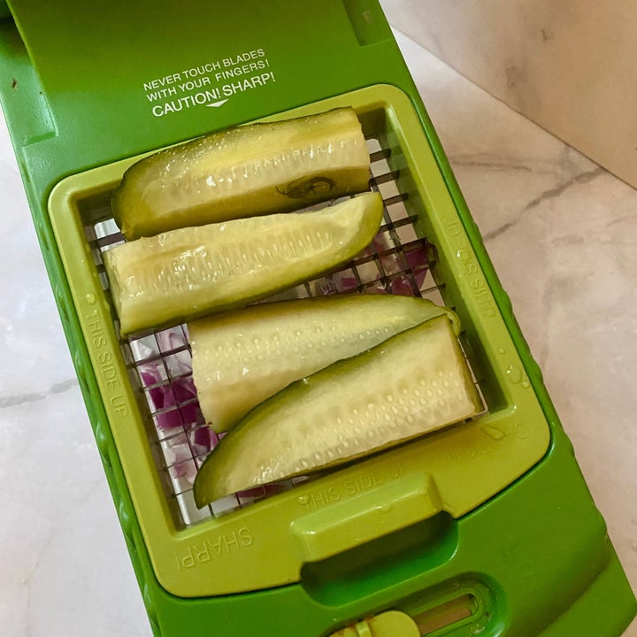 Cucumber slices are placed on a green vegetable slicer with safety instructions visible above the blades