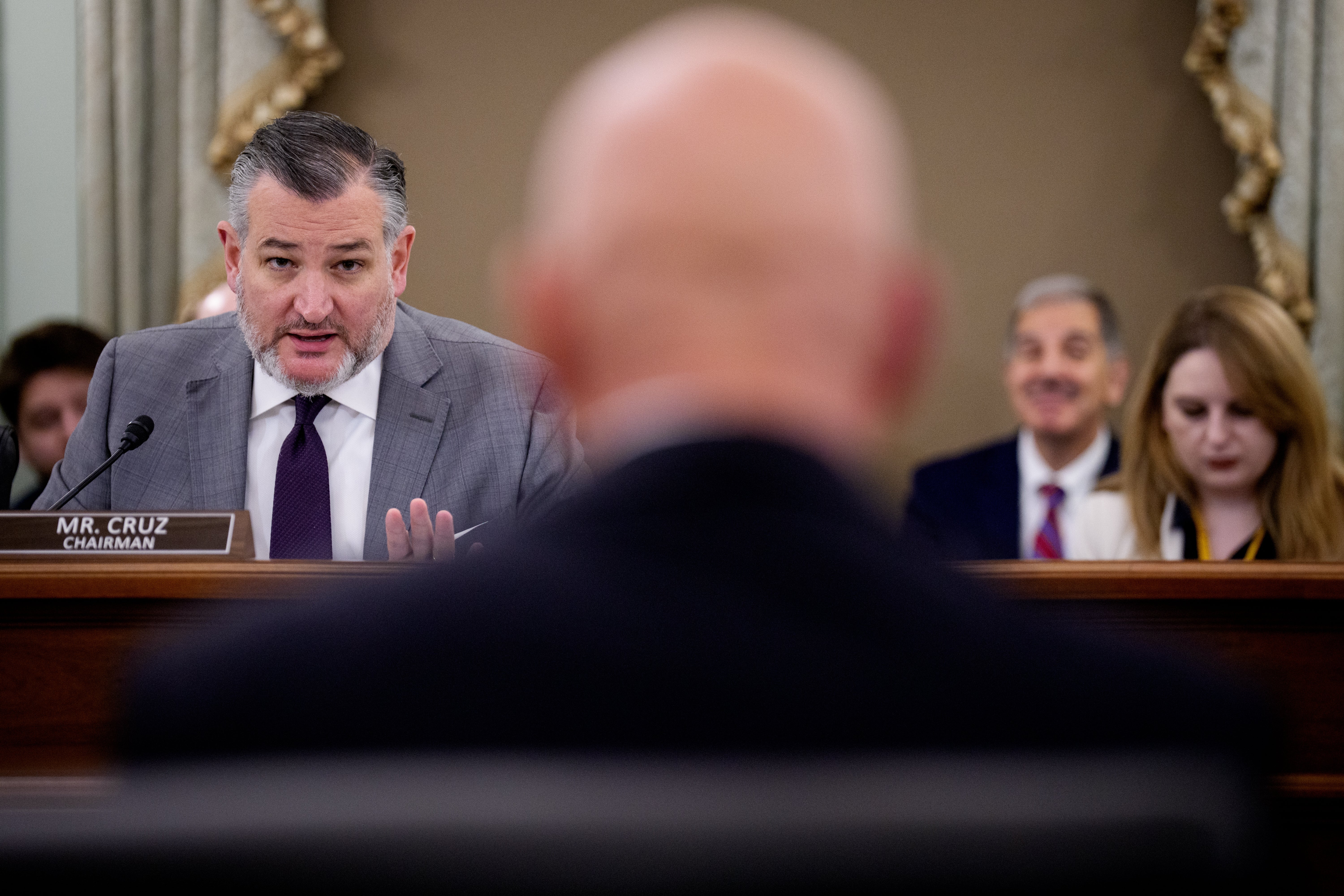 En mann i dress, identifisert som Mr. Cruz, snakker i en mikrofon på et formelt møte, med deltakere synlige i bakgrunnen