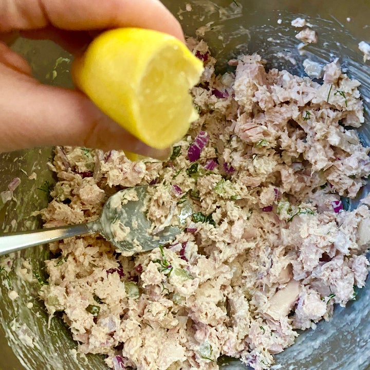 Hand squeezing lemon over a bowl of tuna salad with red onion and herbs, a spoon is in the bowl