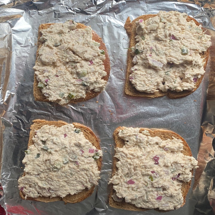 Four slices of bread topped with tuna salad, featuring chunks of onion and celery, laid on a foil-covered surface