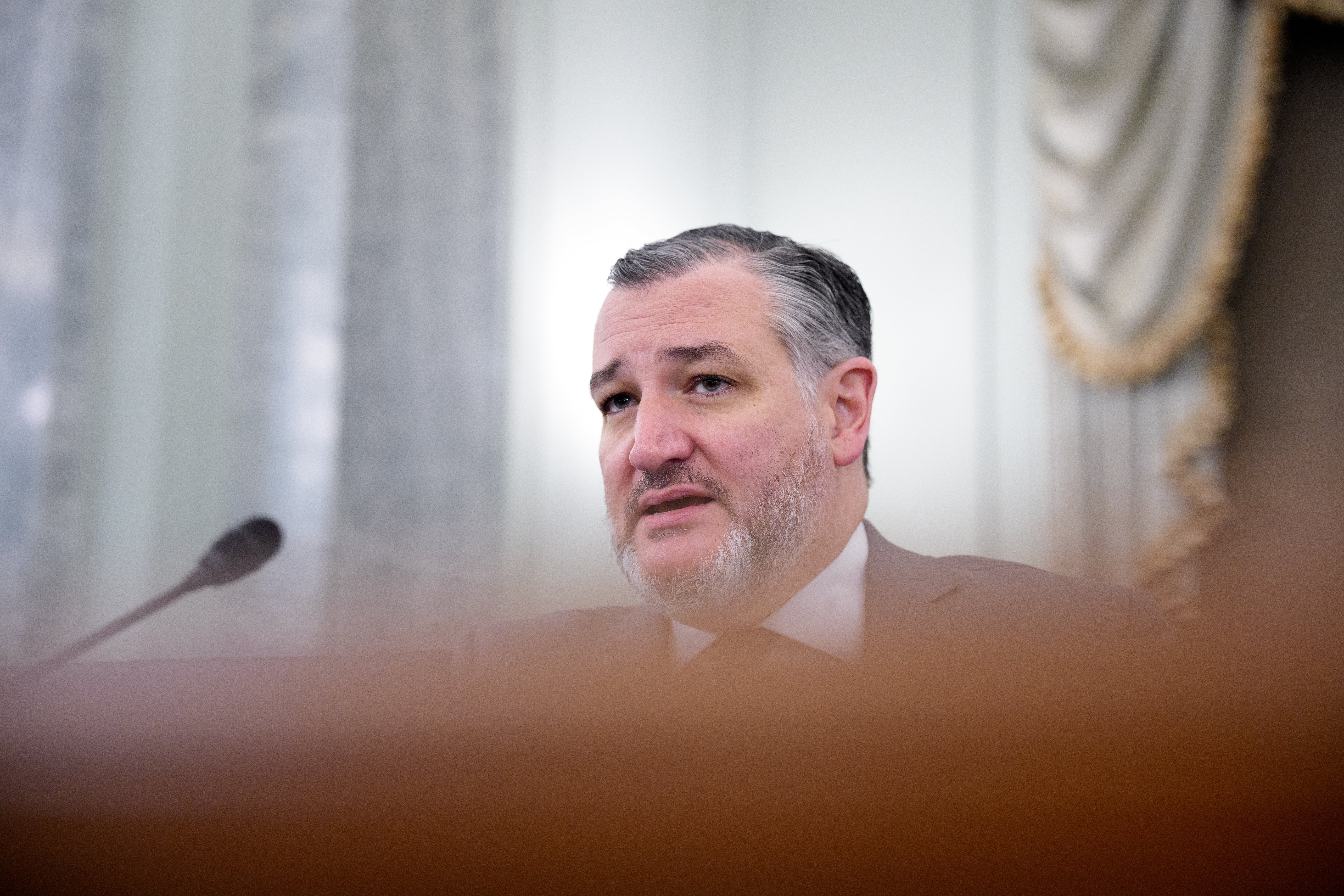 En mann i dress ser ut til å snakke på et formelt arrangement med en mikrofon synlig, gardiner i bakgrunnen