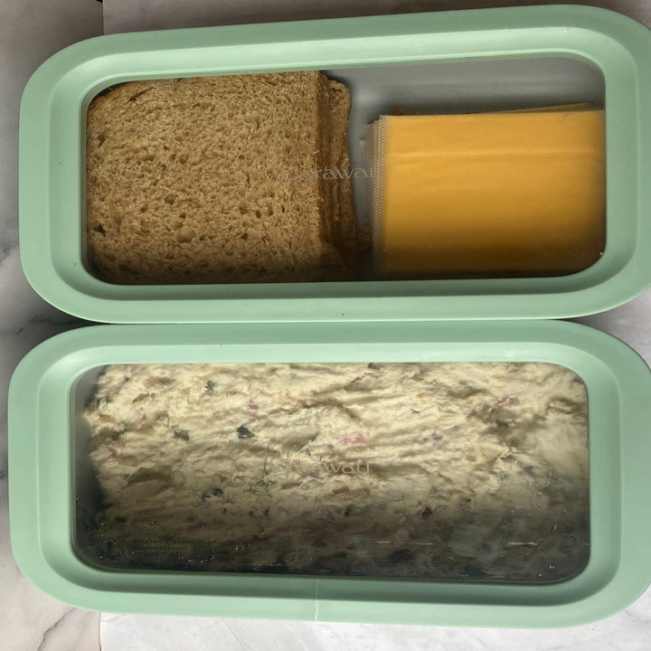 Sandwich container with separate sections for bread, a slice of cheese, and a mixture of salad in the other section. Glasses nearby