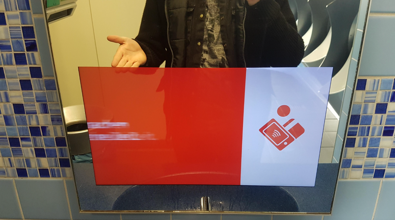 A person stands in front of a bathroom mirror, holding up a hand next to a screen showing contactless payment icons