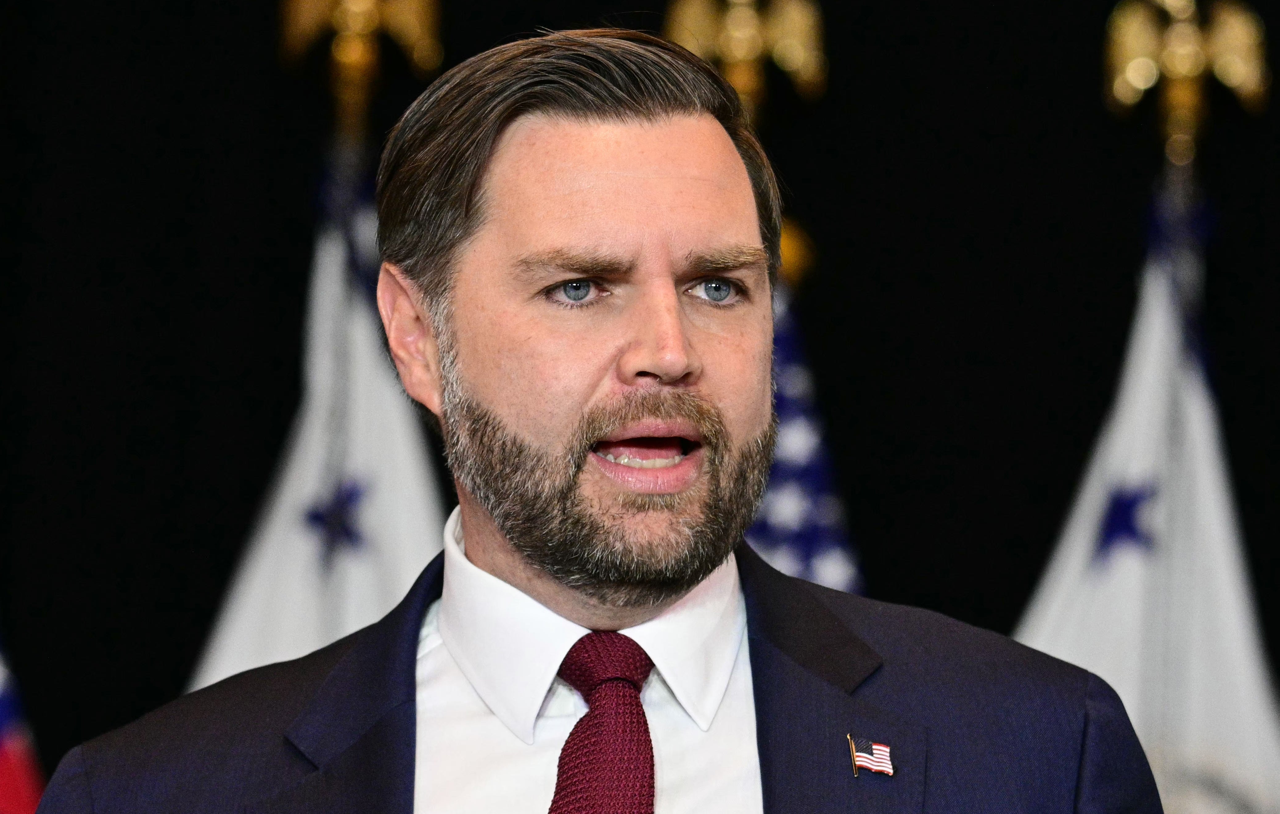 En mann i dress og slips snakker på et arrangement, med flagg i bakgrunnen