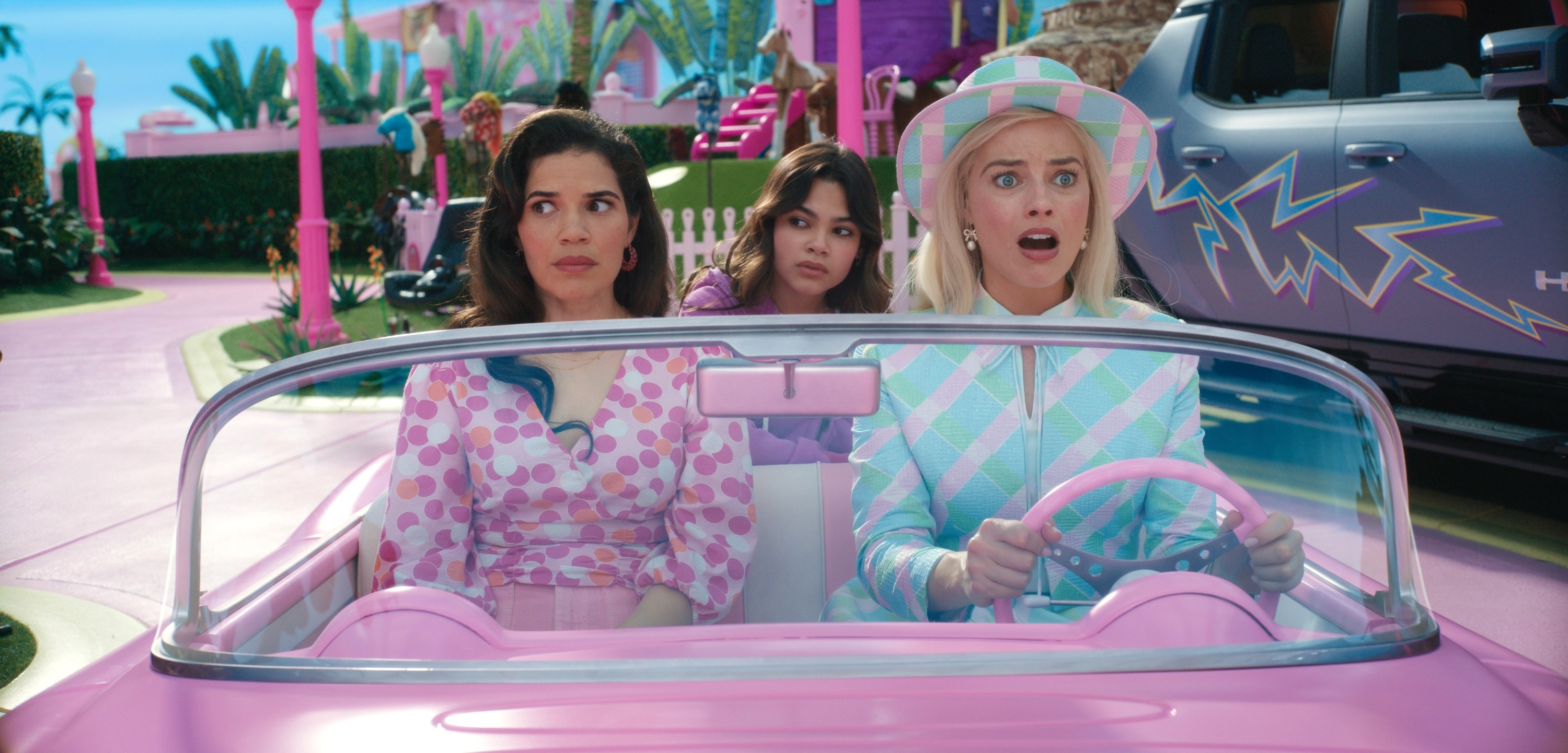 Three people sit in a toy-like convertible car with surprised expressions. They are dressed in colorful, patterned clothing. The background is whimsical
