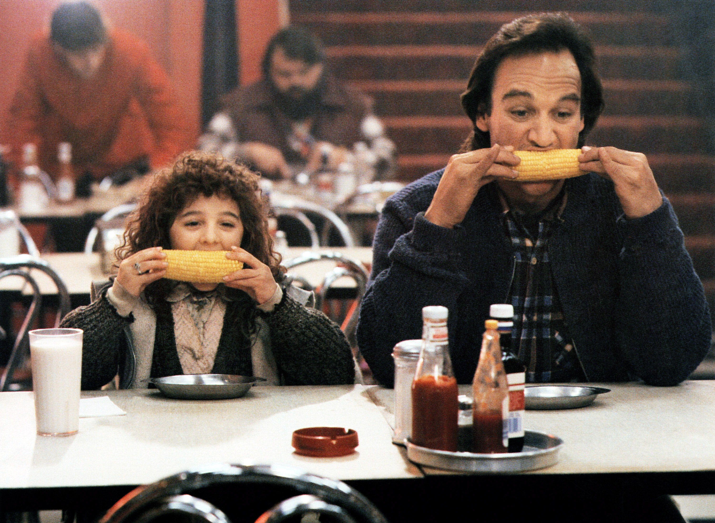 A man and a child sit side by side in a diner, both eating corn on the cob at a table with condiments and a milkshake