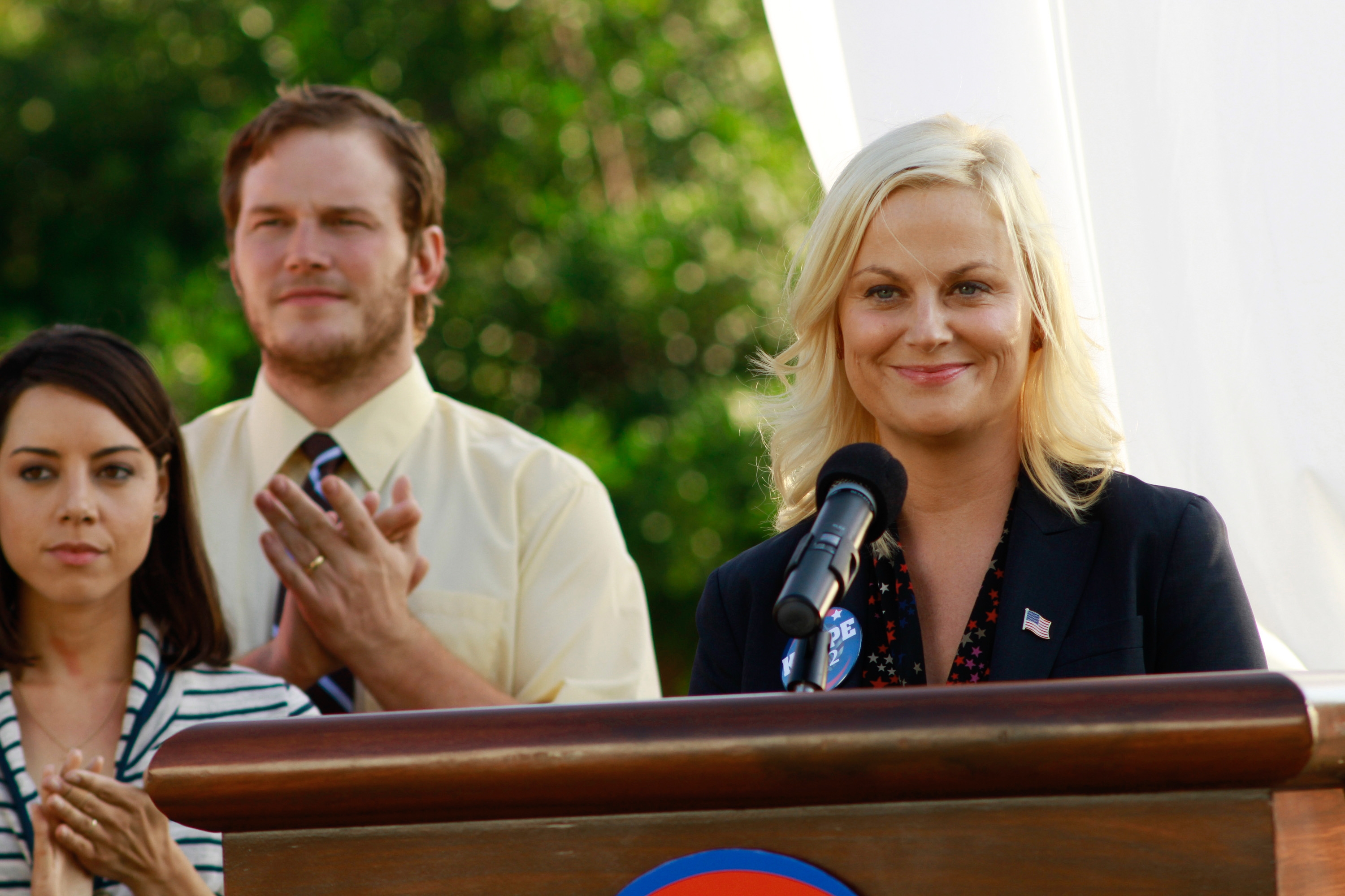 A woman in a blazer speaks at a podium with two people clapping behind her, in an outdoor setting