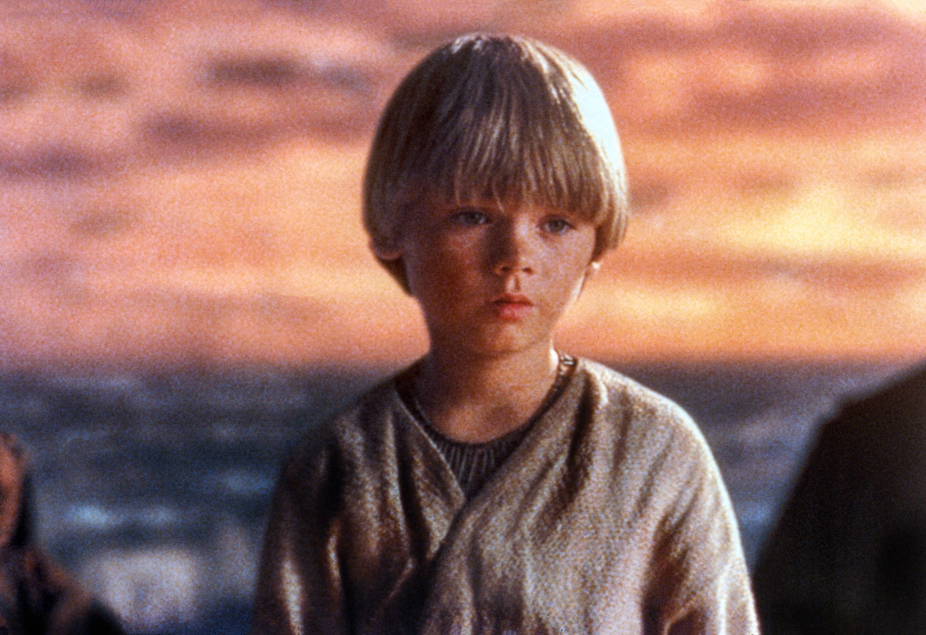 Young child in a robe stands outdoors with a contemplative expression, against a backdrop of sky and water