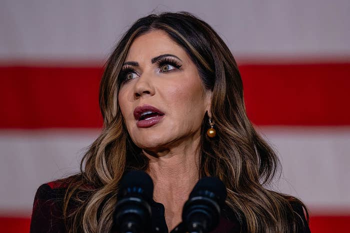 Woman speaking at a podium with microphones, wearing statement earrings, in front of a striped backdrop