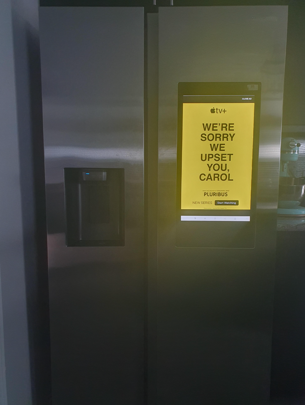 A stainless steel fridge with a screen displaying an ad for Apple TV's new series "We're Sorry We Upset You, Carol."