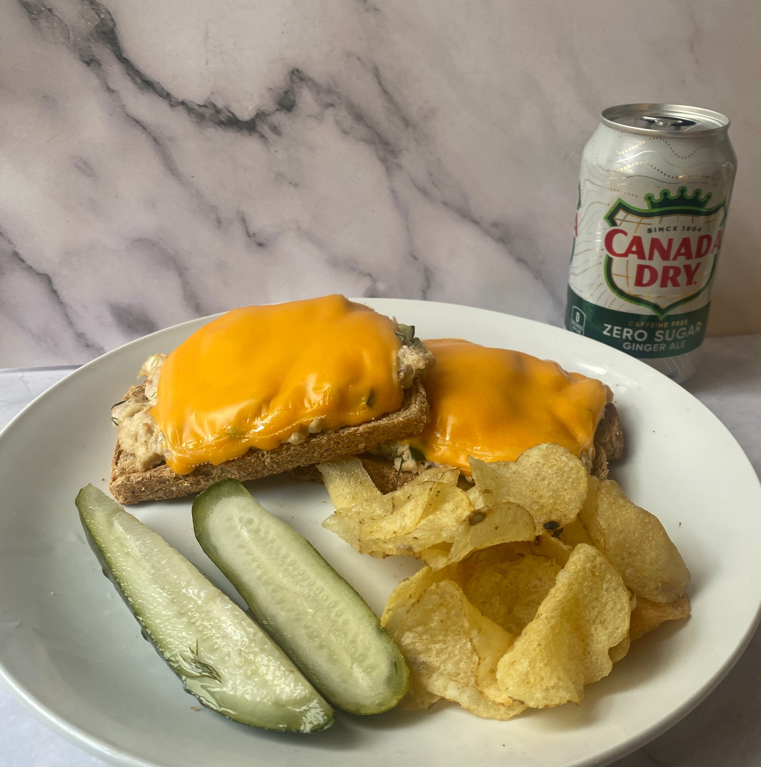Plate with two cheese-covered tuna melts, pickle spears, and potato chips next to a can of Canada Dry ginger ale
