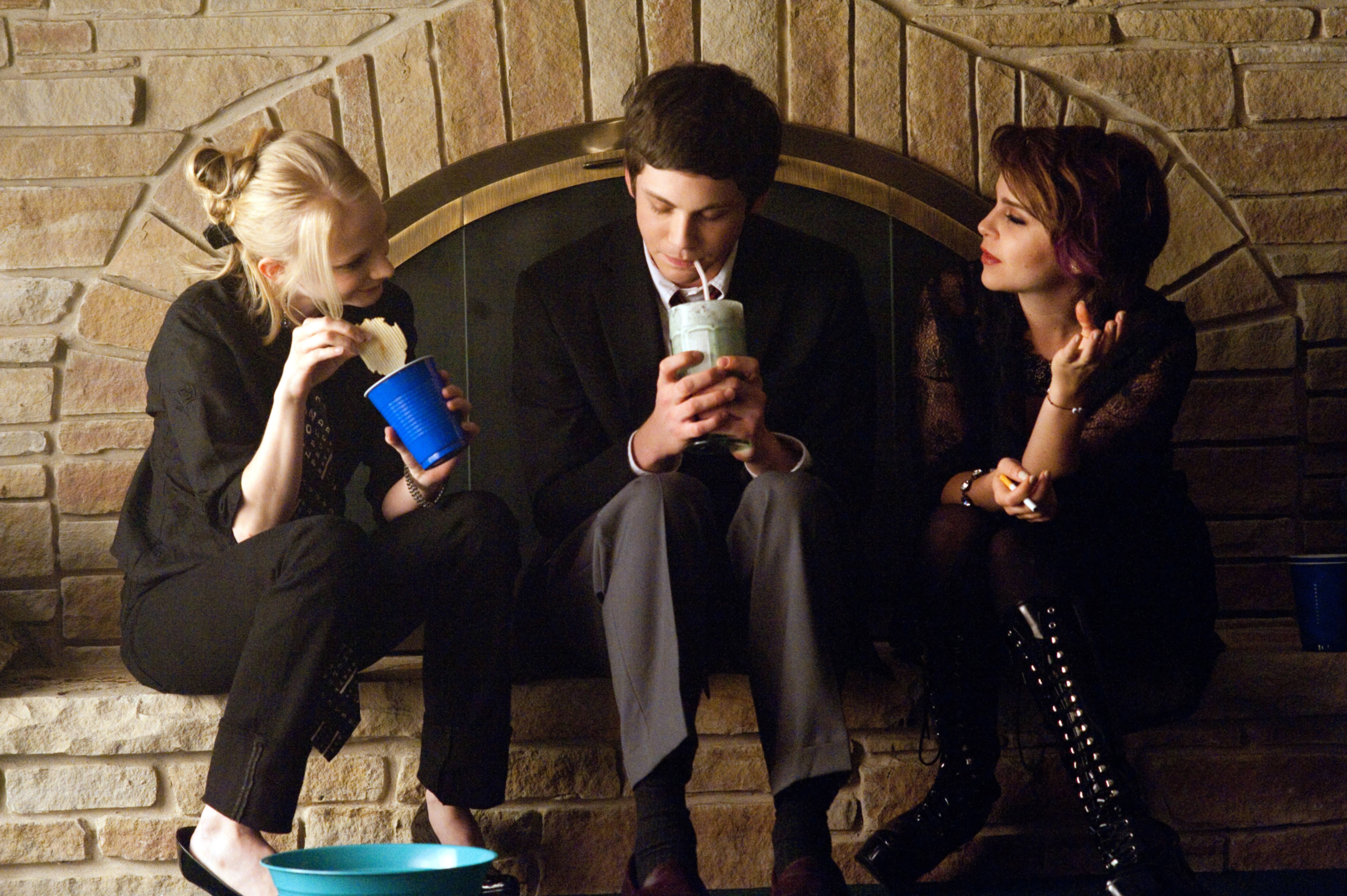 Three people sit by a fireplace. The person in the middle holds a large drink. Casual conversation and relaxed attire
