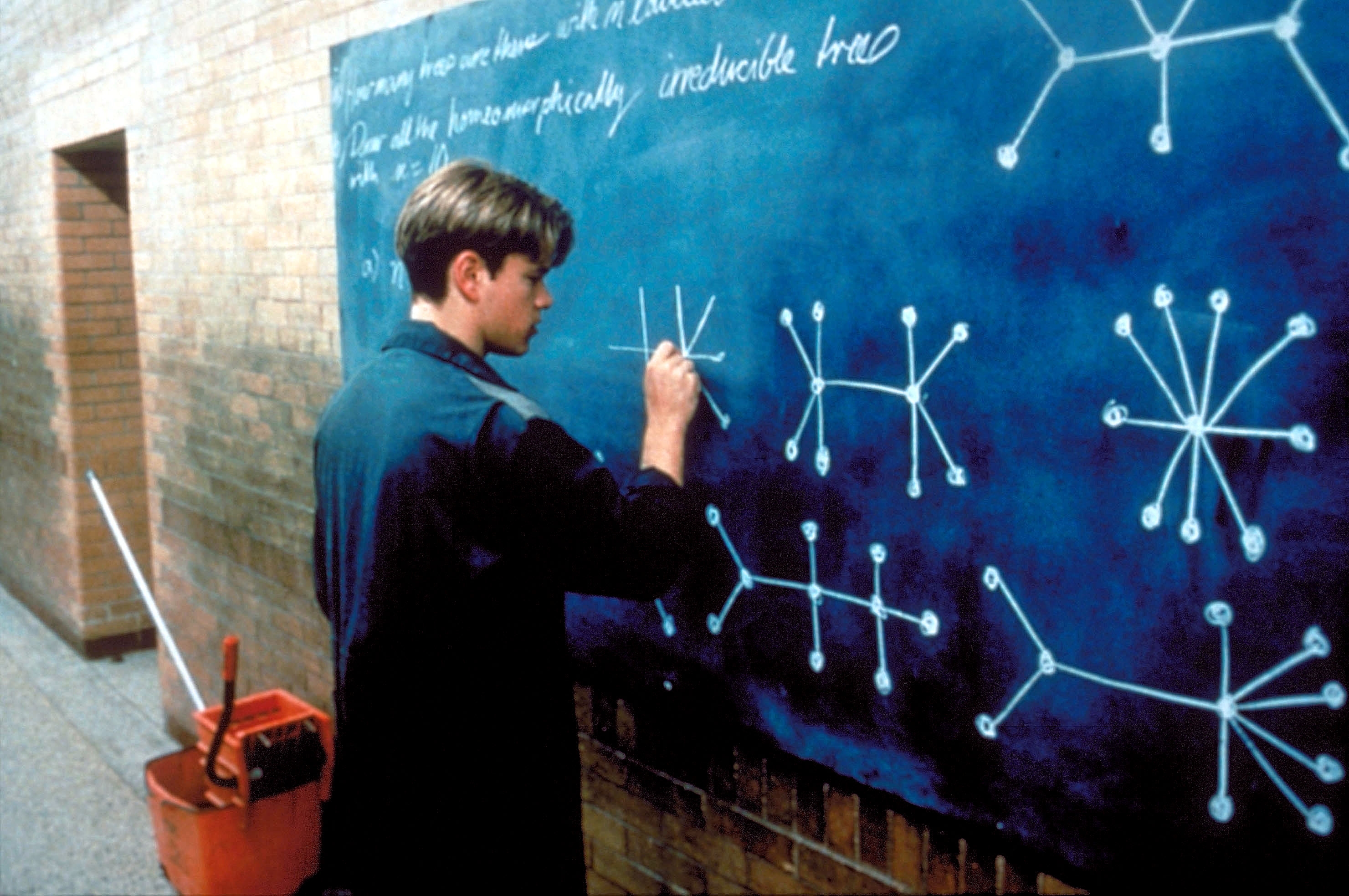 Person writing complex equations on a large chalkboard in a hallway, wearing a casual jacket. A mop and bucket are nearby