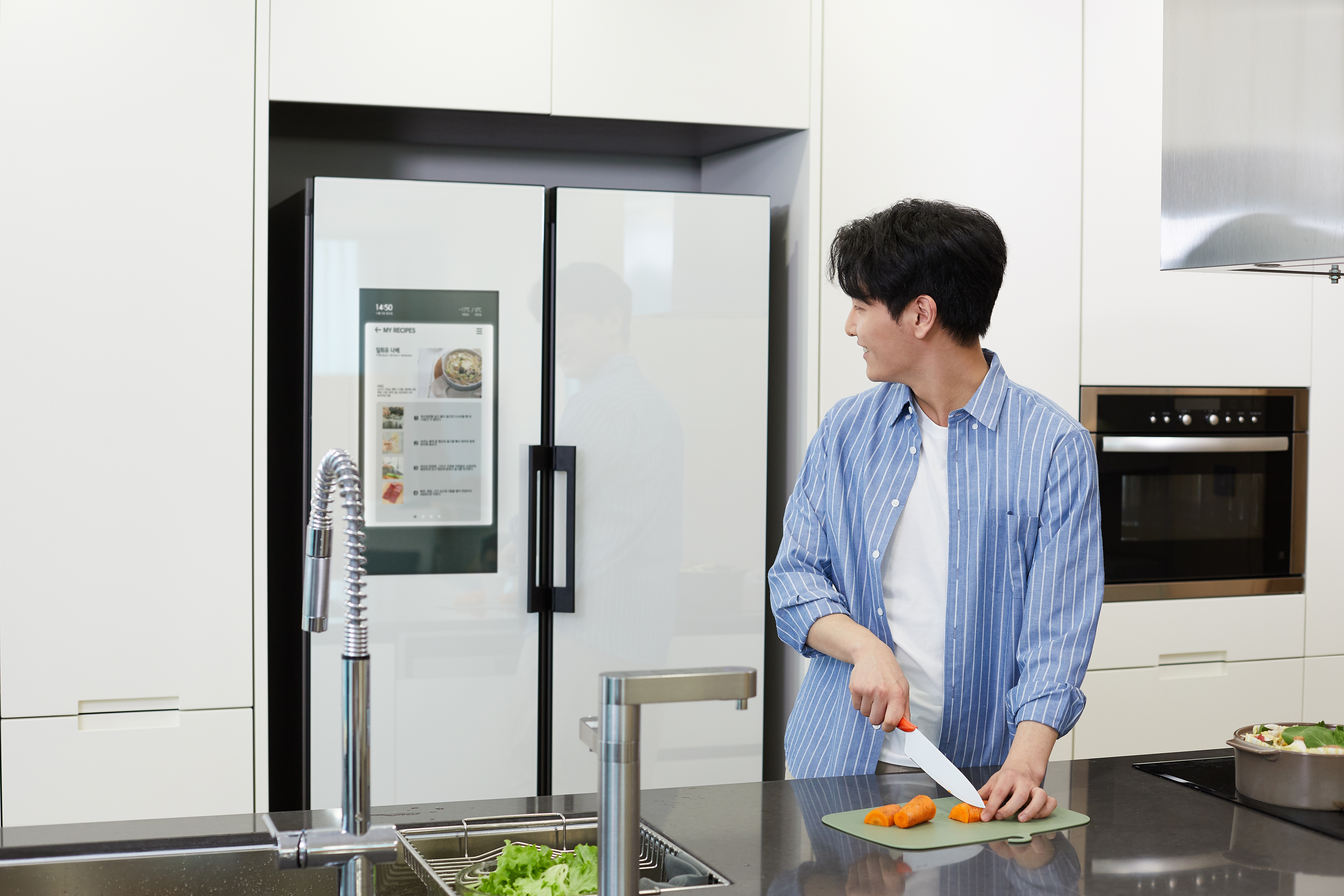 Person som kutter grønnsaker i et moderne kjøkken, ser på et smart kjøleskap som viser en oppskrift