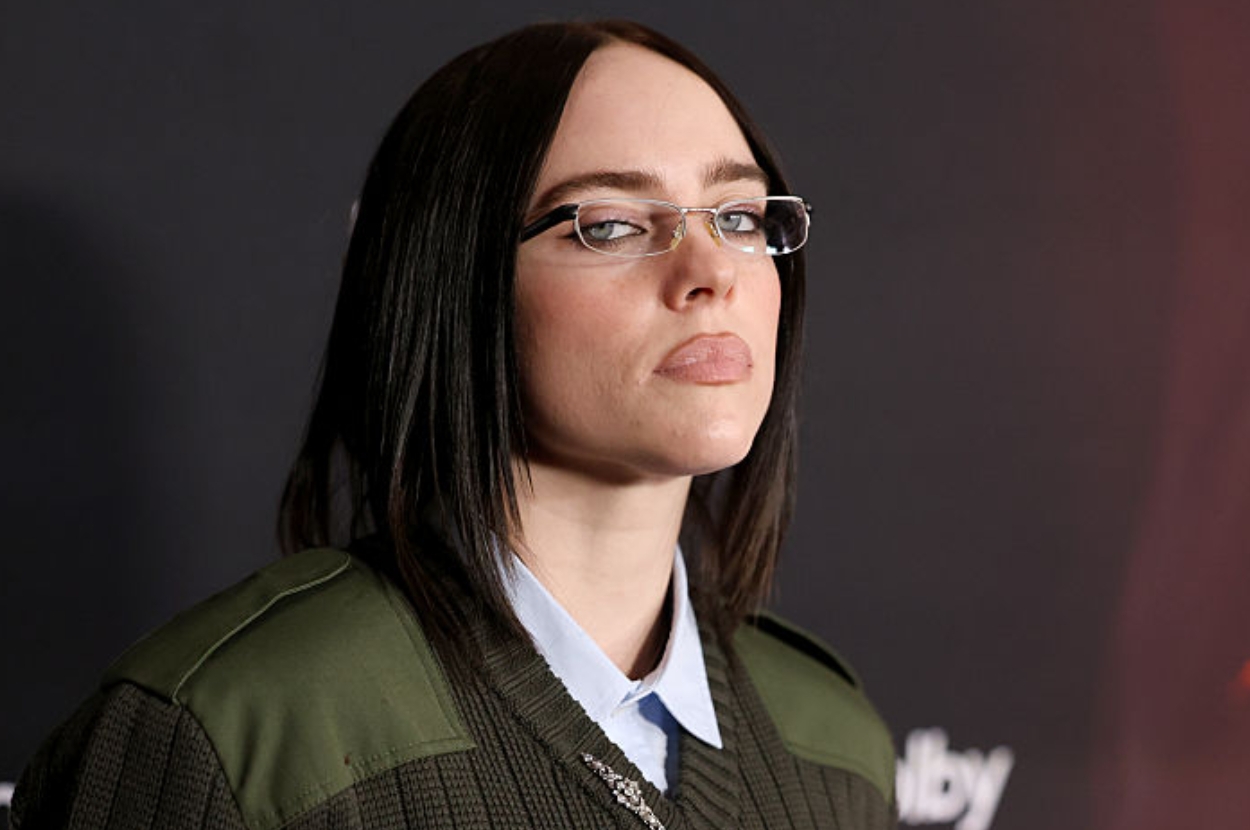 Person with dark hair and glasses wearing a layered outfit, including a collared shirt and a knit sweater, stands against a dark backdrop
