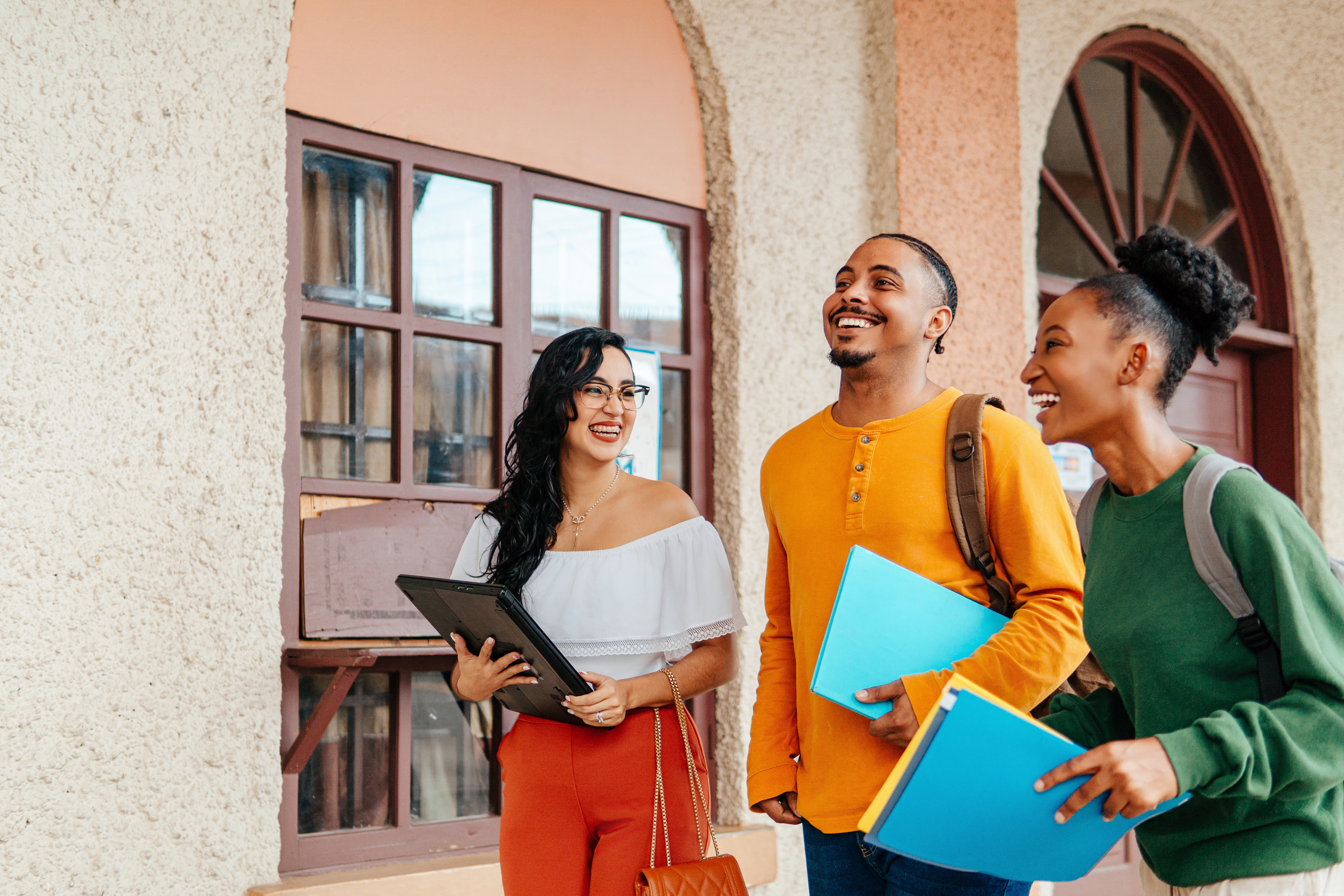 Three radical smiling, holding folders and books, walking extracurricular a gathering with arched windows. Casual attire suggests a pupil setting