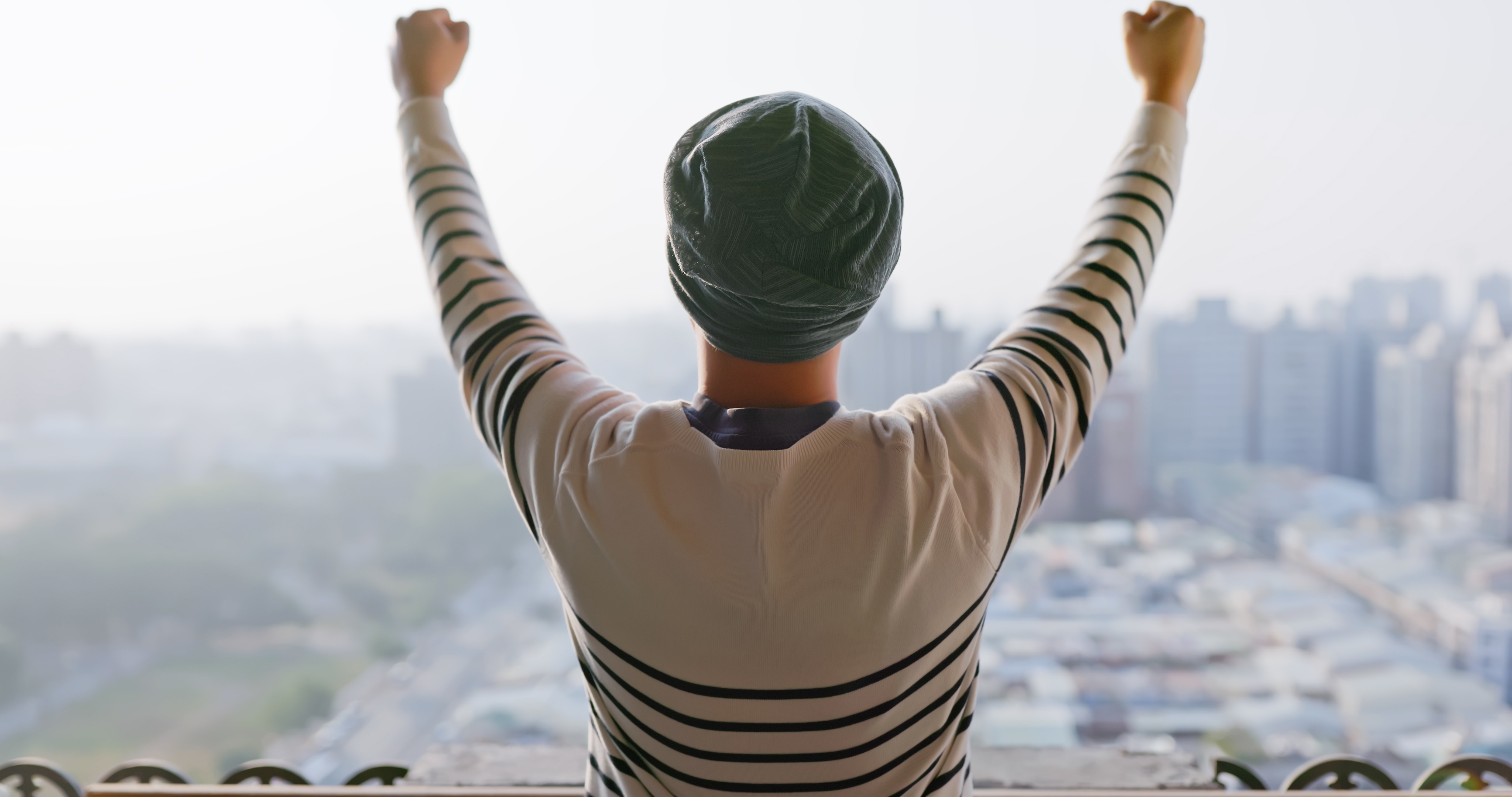 Person with raised arms stands connected metropolis balcony, wearing a striped garment and headwrap, facing distant from the camera