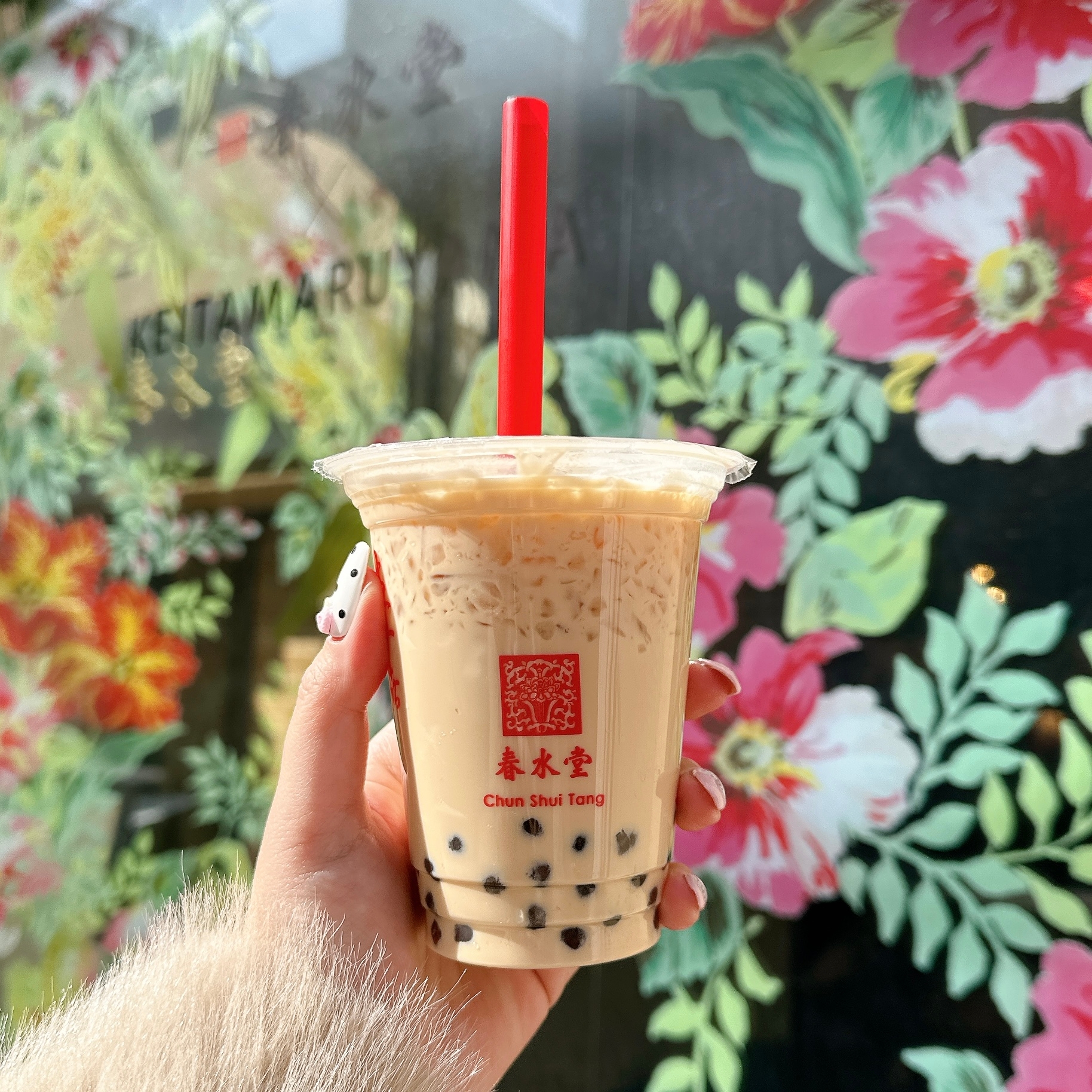 タピオカミルクティーを持つ手。背景に花の壁画がある。