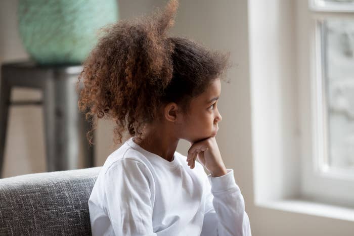 Child with curly hair in a white shirt sitting on a couch, gazing thoughtfully out the window