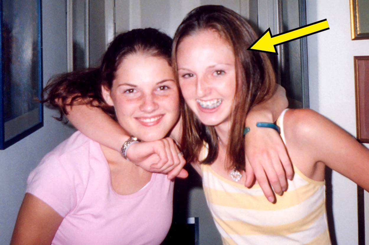 Two smiling young women pose together indoors. The woman on the right has an arrow pointing at her, both dressed casually
