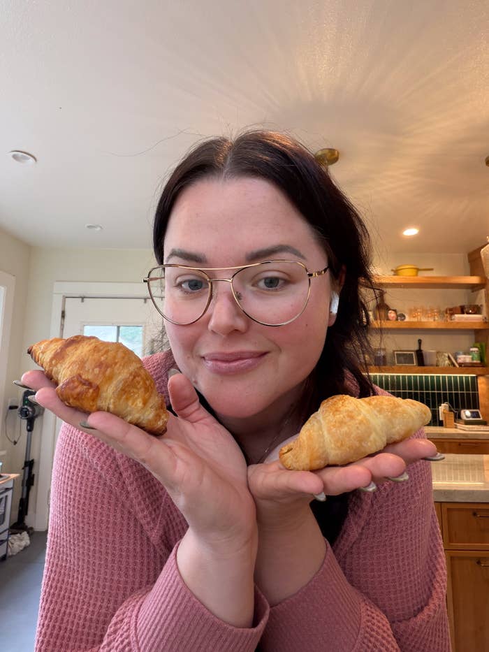 Person in a kitchen smiling and holding two croissants in their hands, wearing glasses and a pink sweater