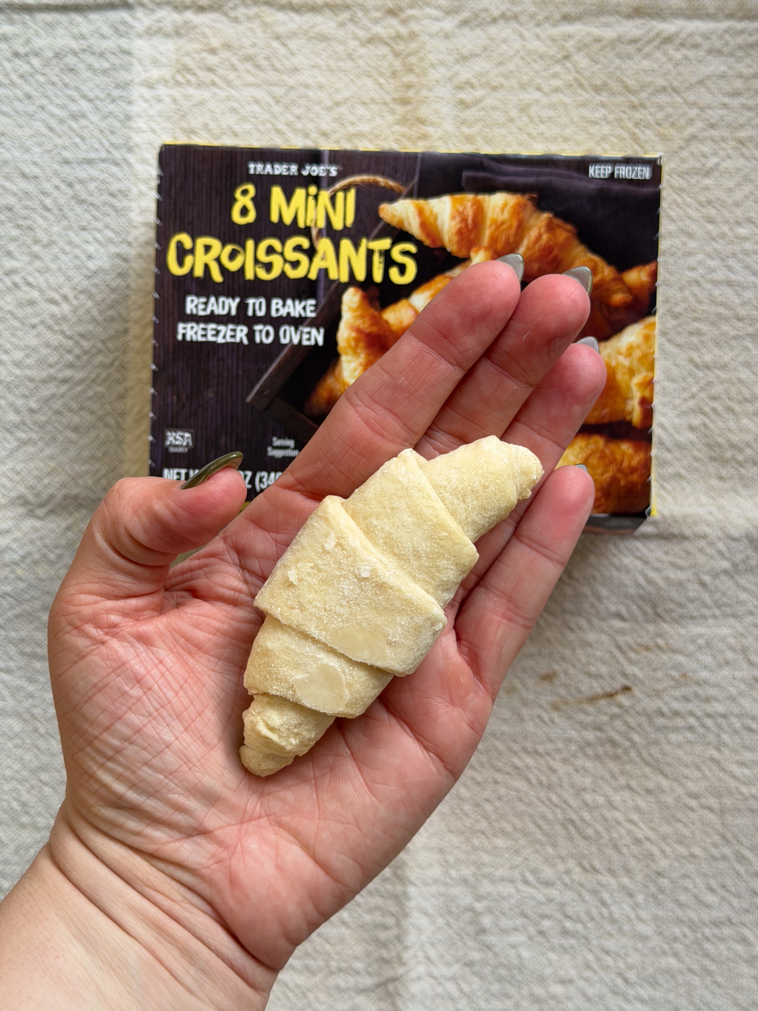 A hand holds an unbaked mini croissant. In the background, a Trader Joe's box advertises "8 Mini Croissants."