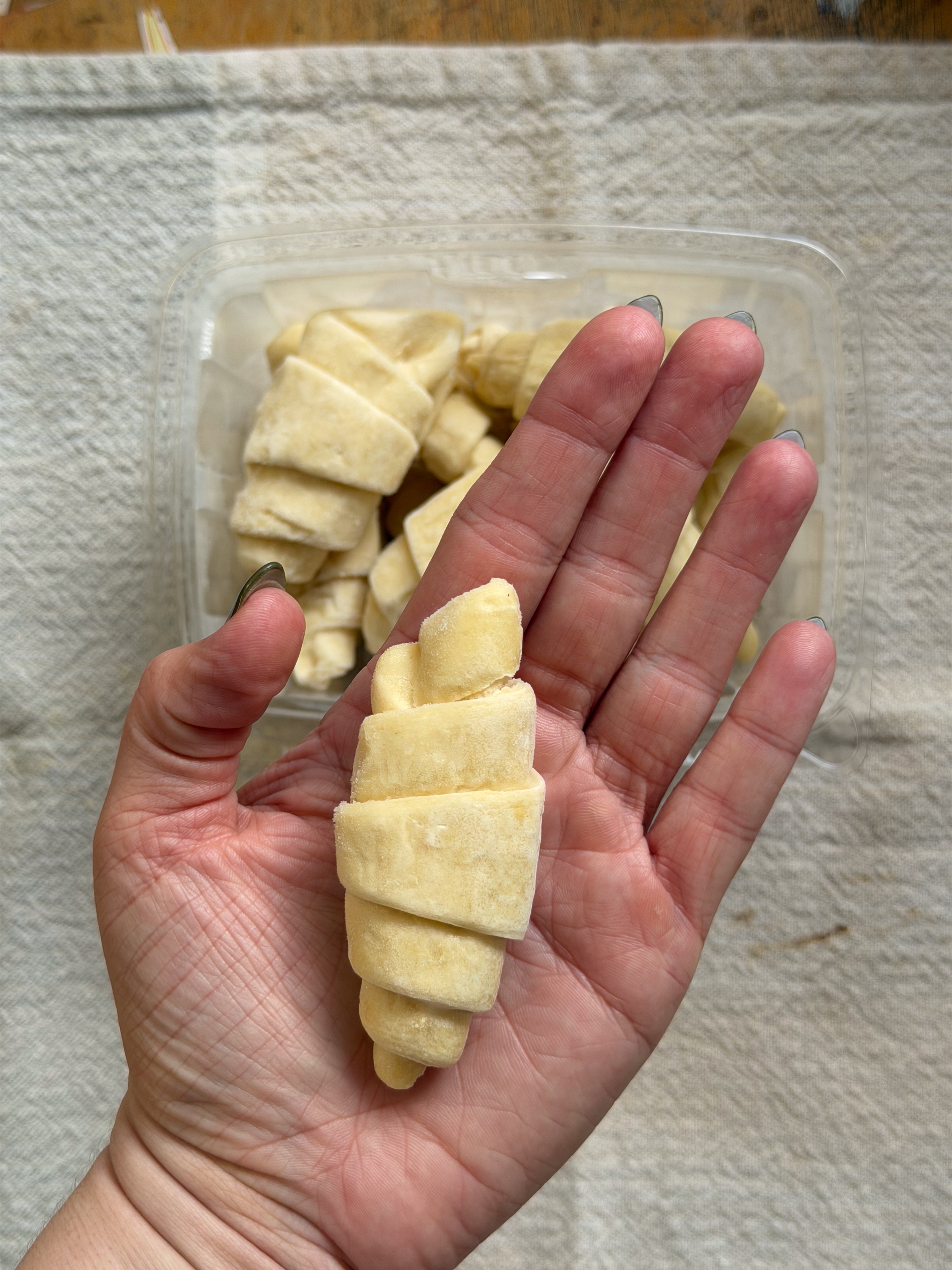 A hand holding a small, uncooked croissant with a tray of croissants in the background on a textured surface