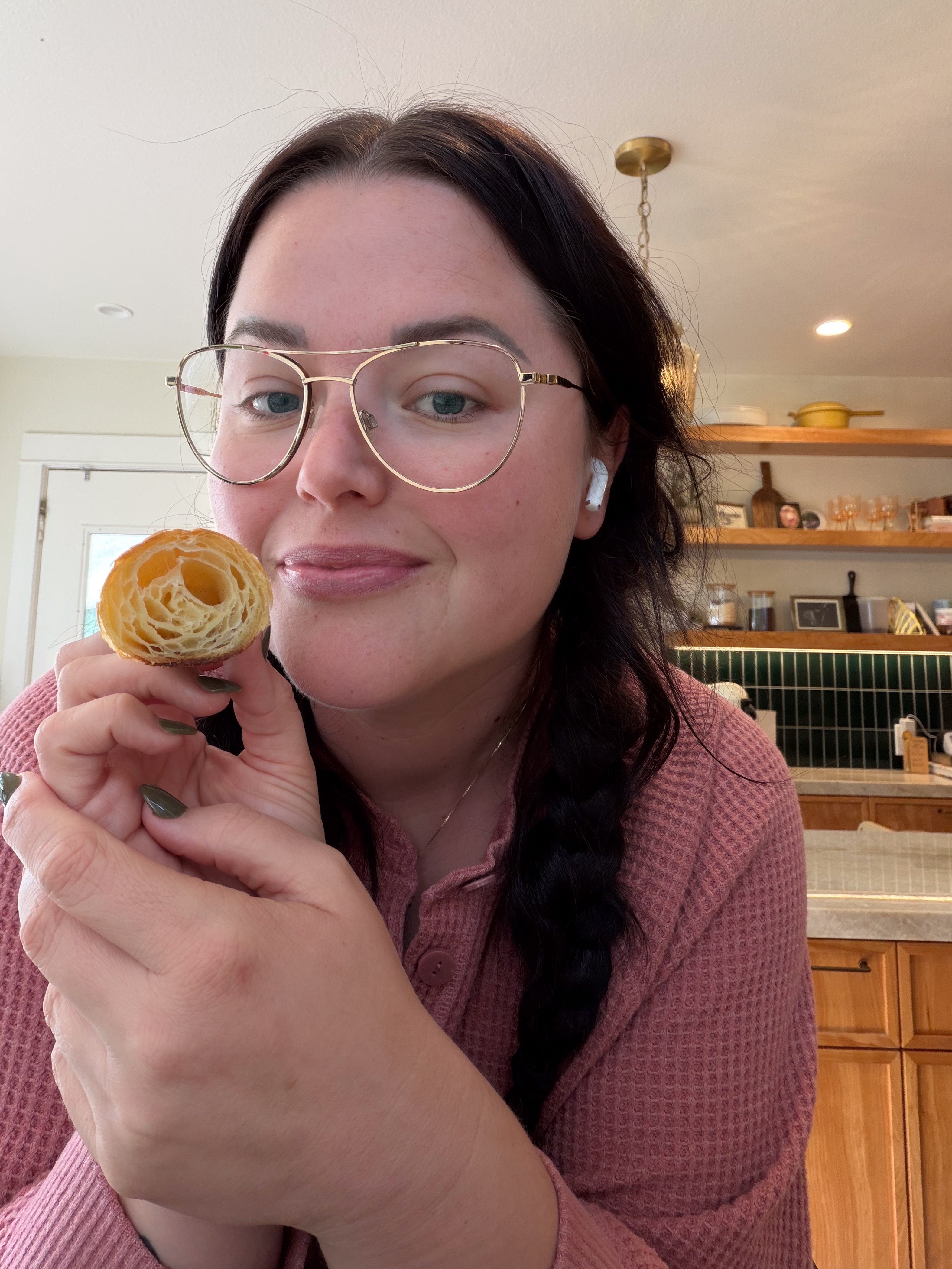Person with glasses holding a sliced pastry, smiling in a kitchen. They have braided hair and are wearing a textured shirt