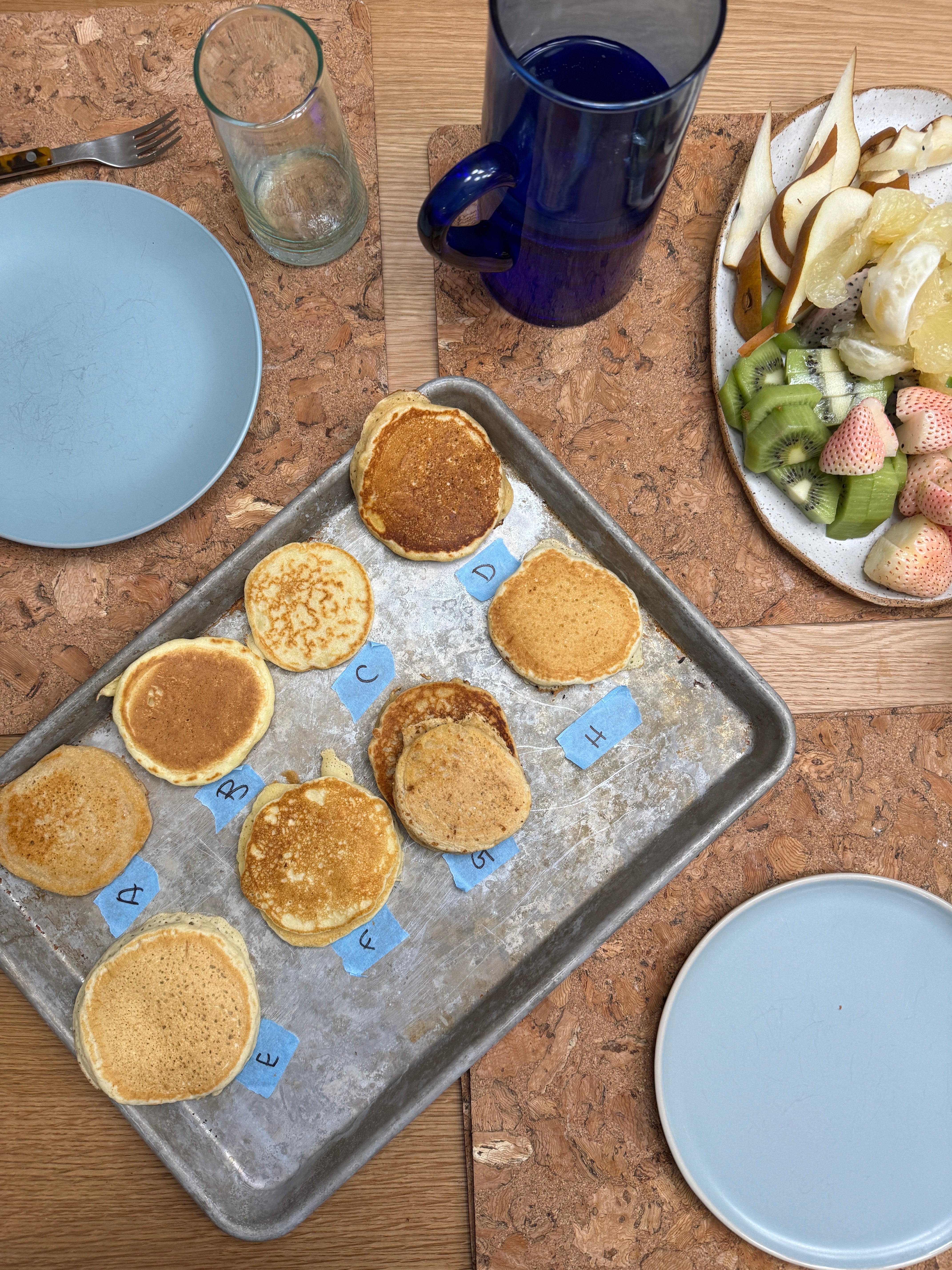 Pancakes labeled A-H on a baking tray, with plates nearby. A platter holds sliced pears, kiwi, and white strawberries. Glass and jug set on cork mats