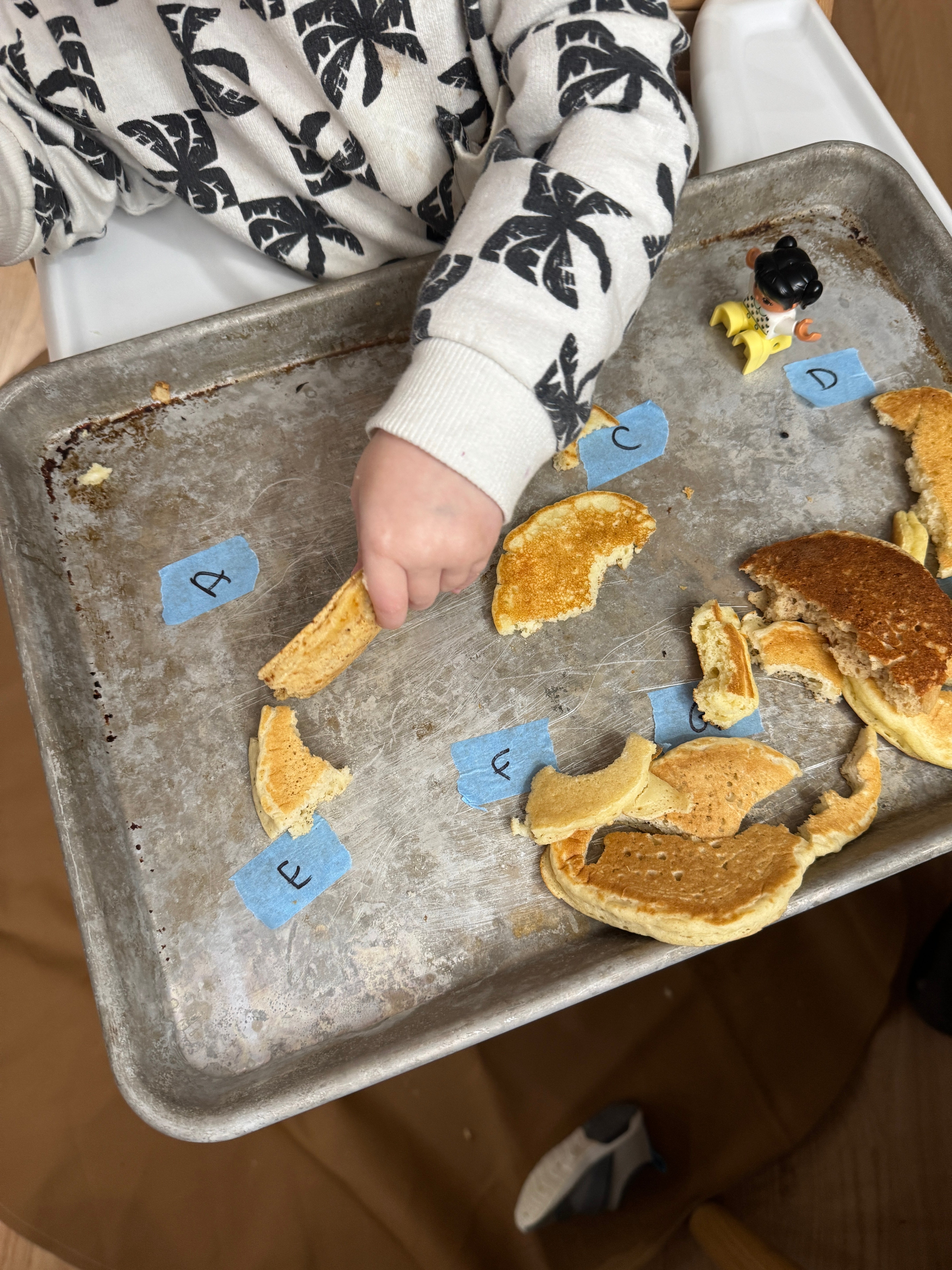 Child wearing a patterned top picks up pancake pieces labeled A-F on a tray