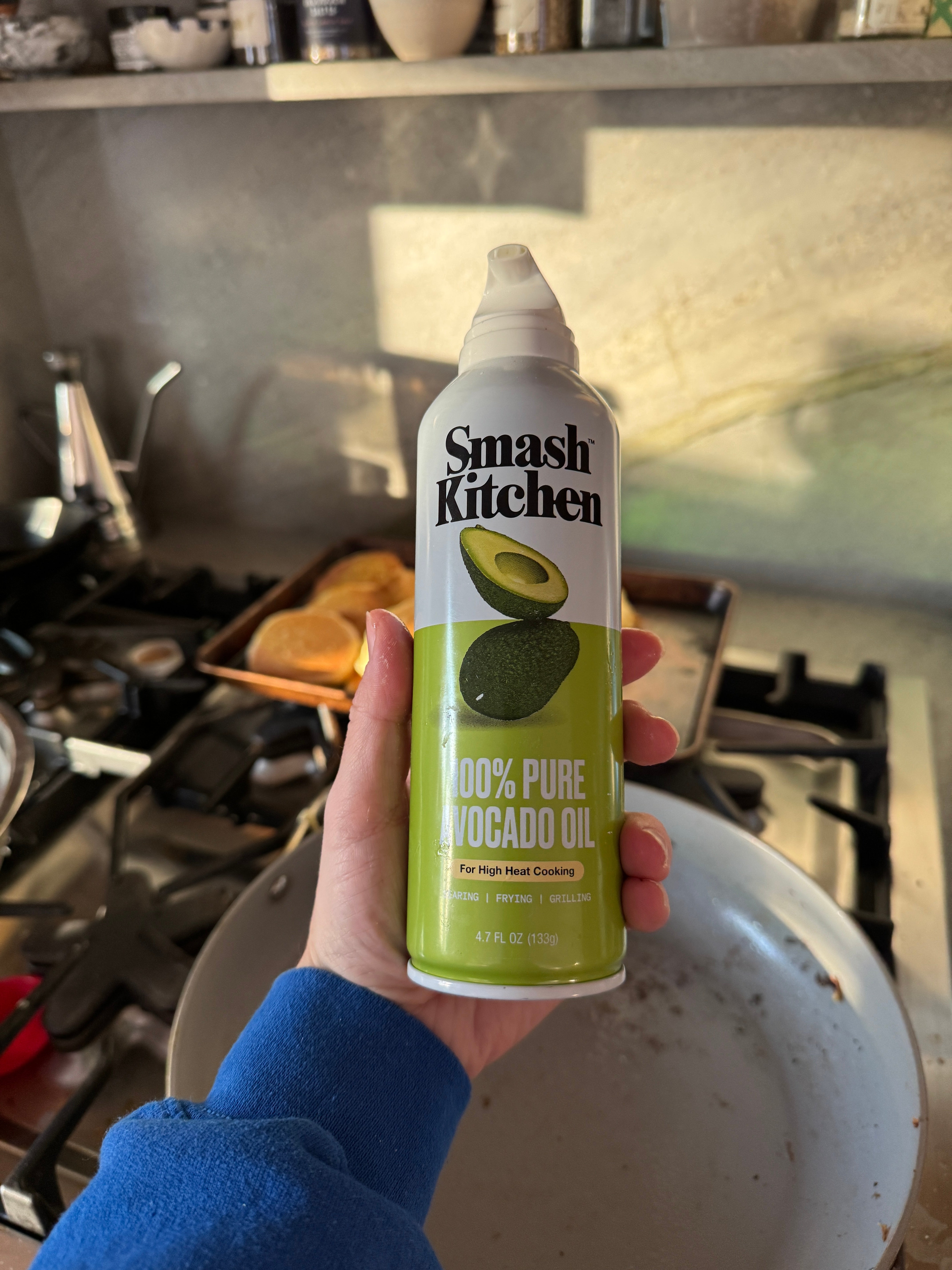 A hand holds a cooking spray can labeled "Smash Kitchen 100% Pure Avocado Oil" in a kitchen setting with cooking utensils in view