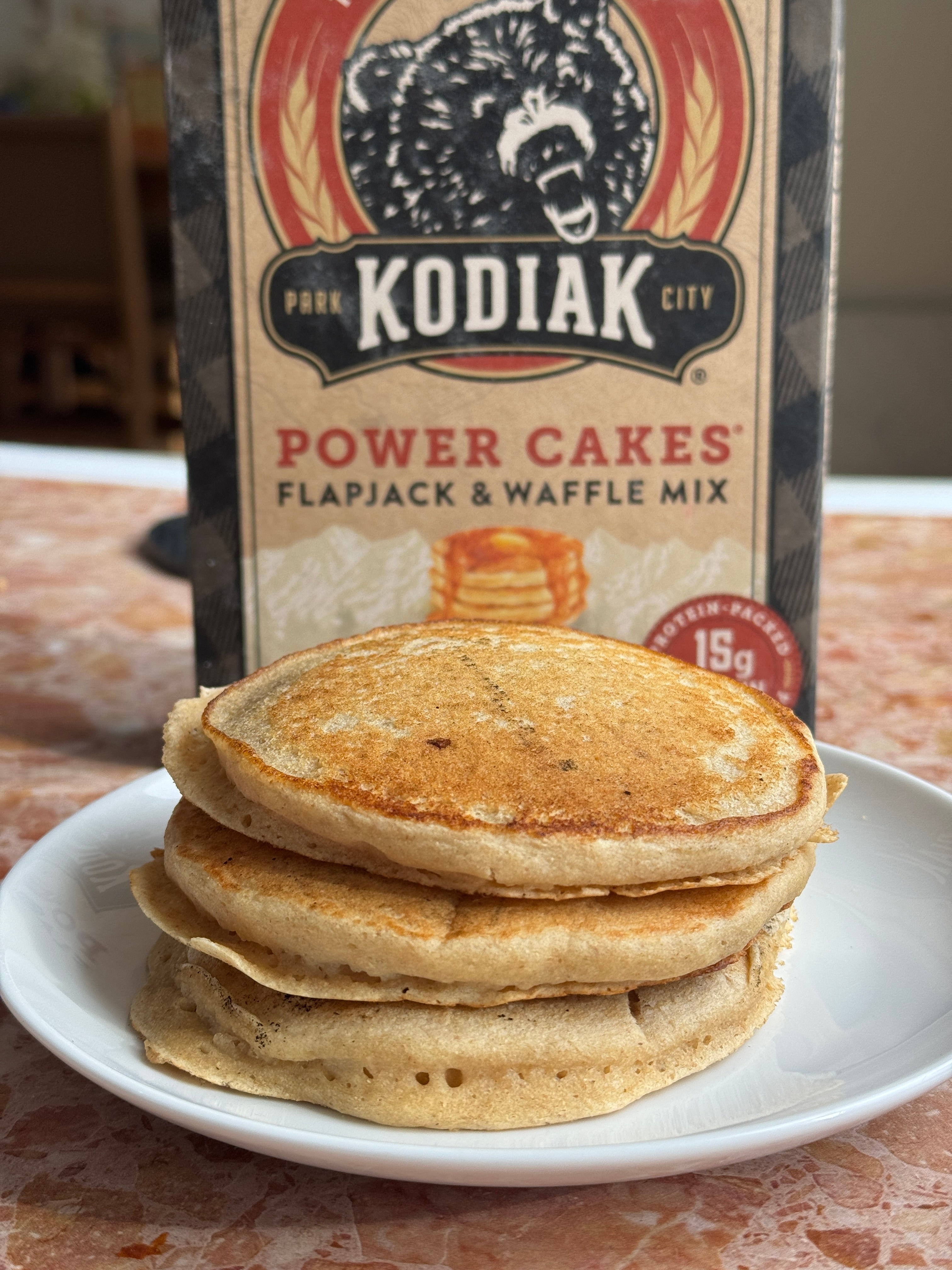 Stack of pancakes on a plate with a box of Kodiak Power Cakes flapjack and waffle mix in the background