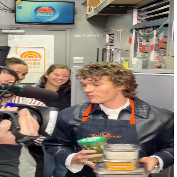 Person in kitchen wearing apron, holding food containers while being filmed by a camera crew. Signs in background suggest a TV show environment