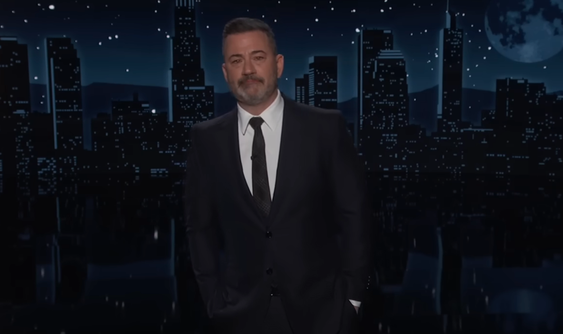 En mann står på en nattlig scene i bybildet i formell dress og slips og holder en tale